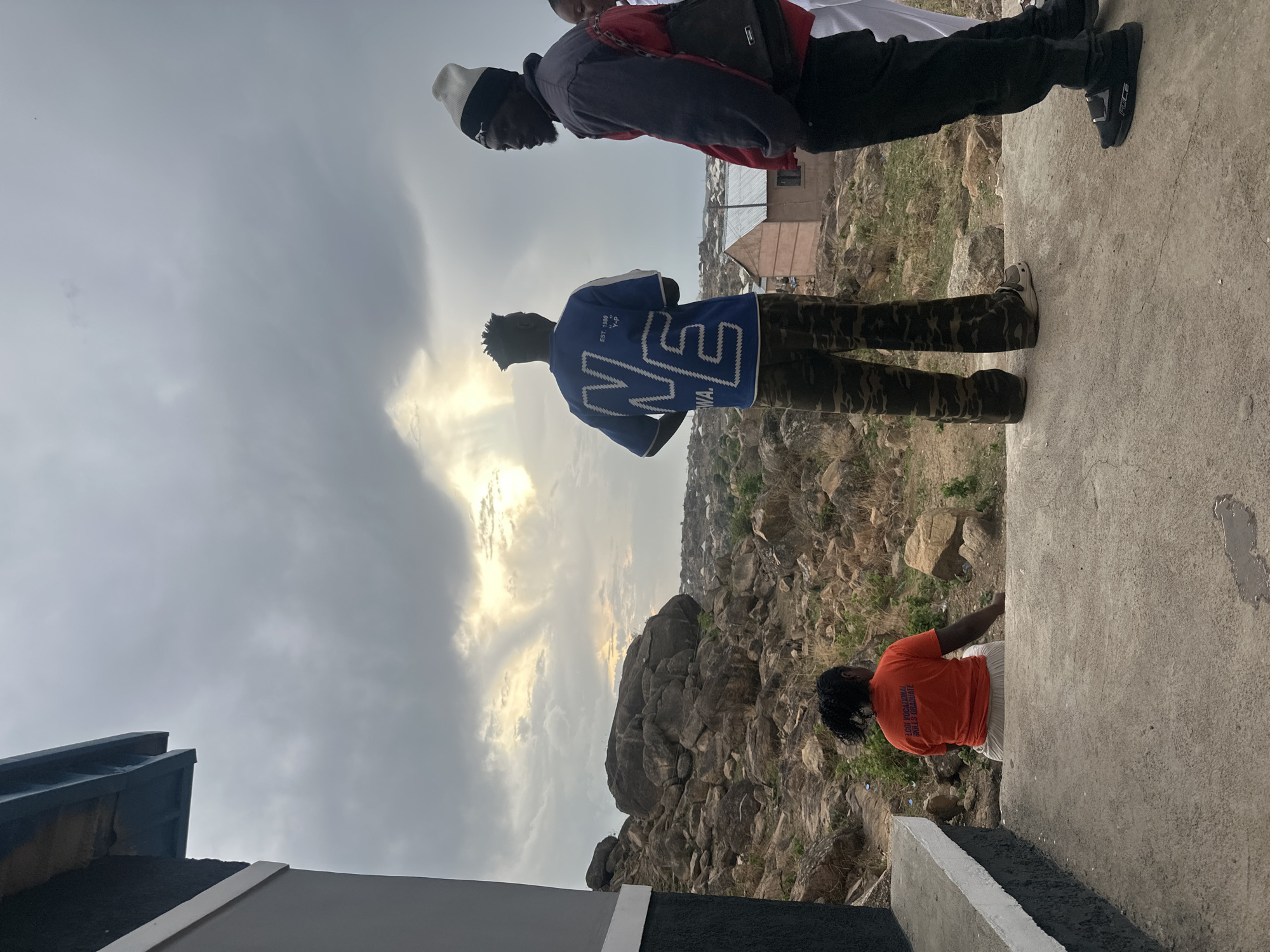 Here is a description of the image:
**Scene:**
The image shows three people standing on a concrete surface. The background consists of a rocky terrain and buildings. The sky is overcast with clouds.
**People:**
* Three young men are visible.
* One man is wearing a blue jersey with camouflage pants.
* Another man is wearing a jacket and a hat.
* The third man is wearing an orange shirt and tan pants.
**Objects:**
* Rocky terrain
* Buildings
* Clouds
* Concrete surface
**Location:**
The image is likely taken in the city of Jos, Nigeria, with the rocky terrain and types of buildings being common in that area.