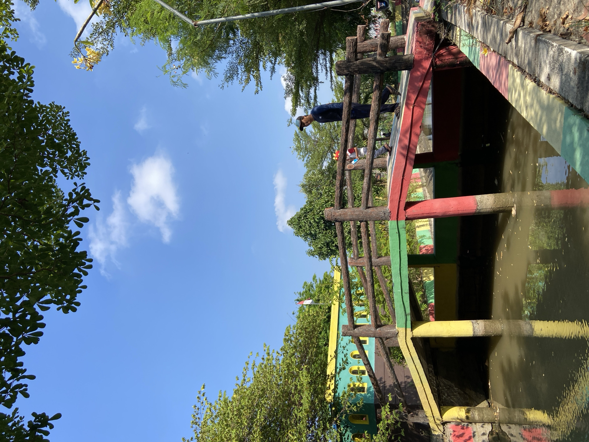 The image depicts a sunny day with a clear blue sky and scattered clouds. In the foreground, a vibrantly painted bridge spans a calm body of water. The bridge features a wooden railing with a rustic texture, and its supporting structure is painted in alternating sections of red, yellow, and green.
On the bridge, a man wearing a dark blue long-sleeved shirt, dark pants, and sandals is walking towards the right side of the frame. He is looking downwards and to his left. Behind him, a child in a red and white shirt and patterned pants is also walking on the bridge. The child is holding a small, light-colored object.
To the left of the bridge, a colorful building with green walls and yellow-framed windows is visible. A red and white flag, likely the Indonesian flag, is seen flying from a pole behind the building. Lush green trees surround the area, with some branches of a large tree hanging from the top of the frame. The overall environment suggests an outdoor recreational space or park. The scene appears to be located in the city of Gelam, Indonesia, given the context.
