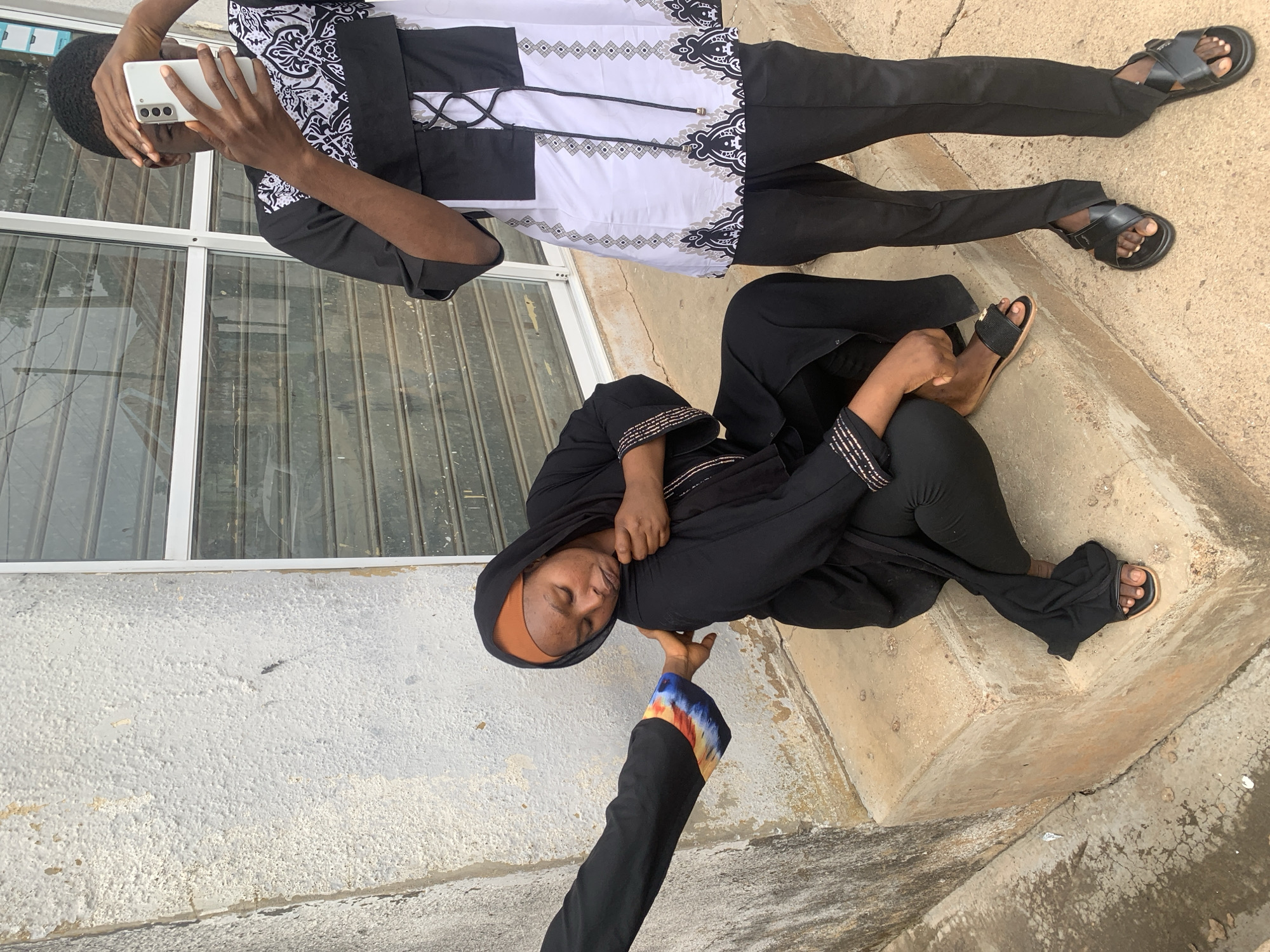 The scene takes place outdoors in the city of Jos, Nigeria, against a textured, peeling white building wall with a roll-down metal shutter window.
A dark-skinned woman, wearing a black hijab over an orange headscarf, a black long-sleeved garment with embroidered cuffs, black leggings, and black flat sandals, is seated on a discolored concrete step. Her eyes are closed, and her expression suggests discomfort or exhaustion. An arm, extending from the left side of the frame and clad in a black sleeve with a distinct multicolored patterned cuff, reaches out to touch the woman's back, appearing to offer support.
To the right, a dark-skinned man stands on the concrete ground. He wears a black and white patterned tunic shirt with a lace-up front, black trousers with ankle slits, and black open-toed sandals. He holds a white smartphone vertically with both hands, obscuring his face as he looks at the screen or records the scene. The concrete ground and steps are worn and display cracks.