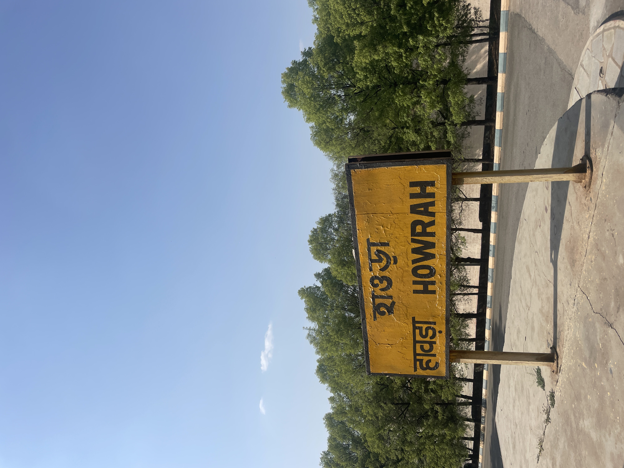 The image depicts a yellow signpost with the word "HOWRAH" written in English and Hindi. The signpost is located on a concrete surface near a tree-lined road, under a clear blue sky. It marks the location of Howrah, a city near Anajpur, India.