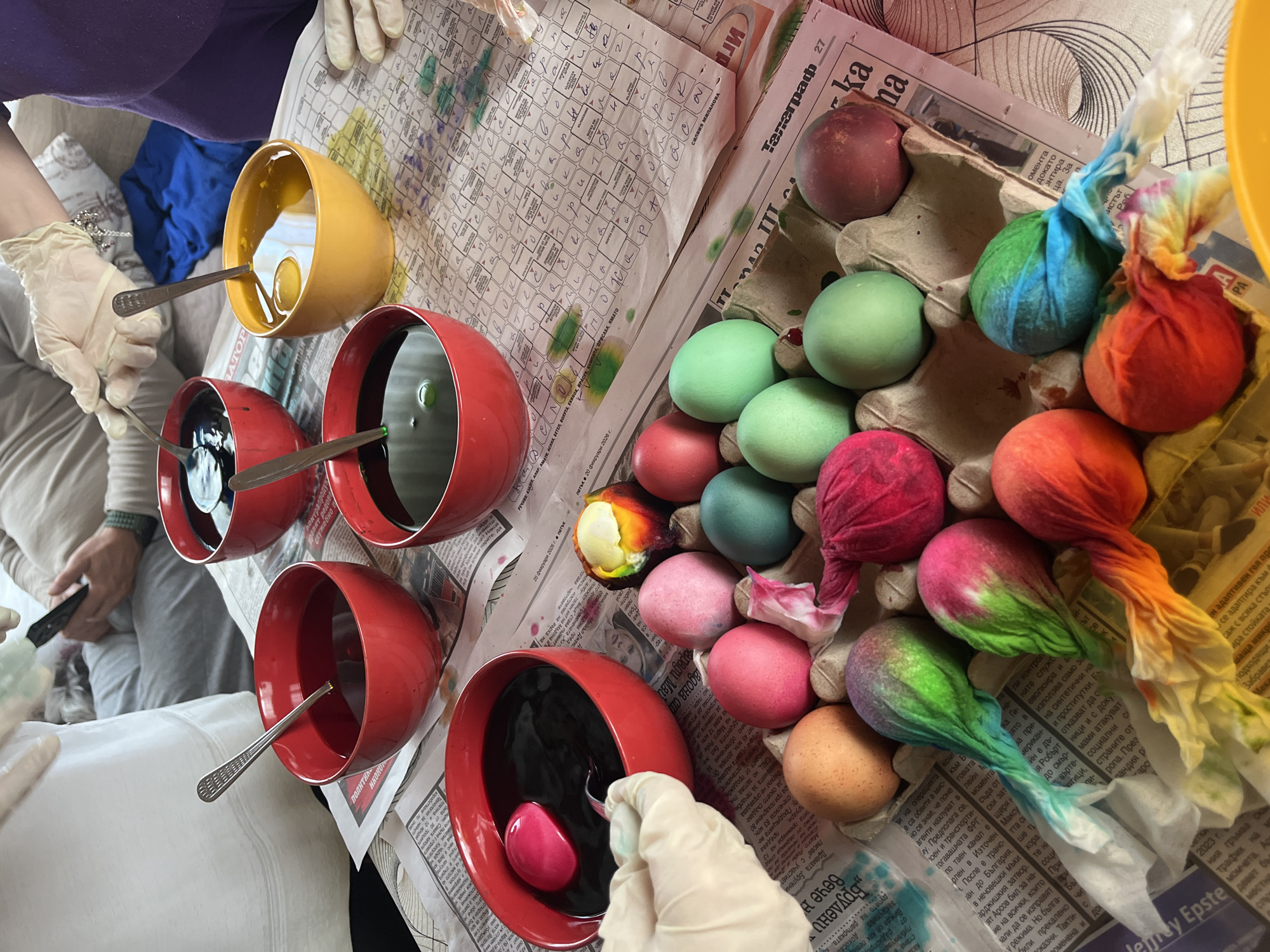 A collection of dyed Easter eggs and dyeing paraphernalia is arranged on a newspaper-covered surface. Several red bowls and one yellow bowl contain liquid dye. Spoons are in most of the bowls. Several eggs, dyed in various colors and some tied with fabric scraps, sit in an egg carton. One hand, wearing a latex glove, appears to be dipping an egg into a bowl of dark dye. Another hand holds a black object. The newspaper is in Cyrillic.