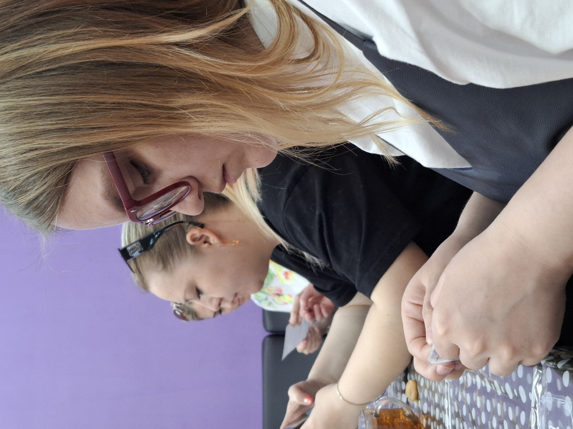 The image depicts three women sitting at a table covered with a patterned tablecloth. The woman in the foreground has blonde hair, wears eyeglasses with red frames, and is dressed in a white top with a dark vest. She is looking down, seemingly focused on something on the table. To her right is another woman with blonde hair pulled back, wearing sunglasses on her head and a black top. She is also looking down at something, holding a small white card in her hands. Partially visible behind her is a third person. The background consists of a solid purple wall. A glass of dark liquid is visible on the table. The location is Markovskii, Russia. The people are engaged in an activity involving small white cards.