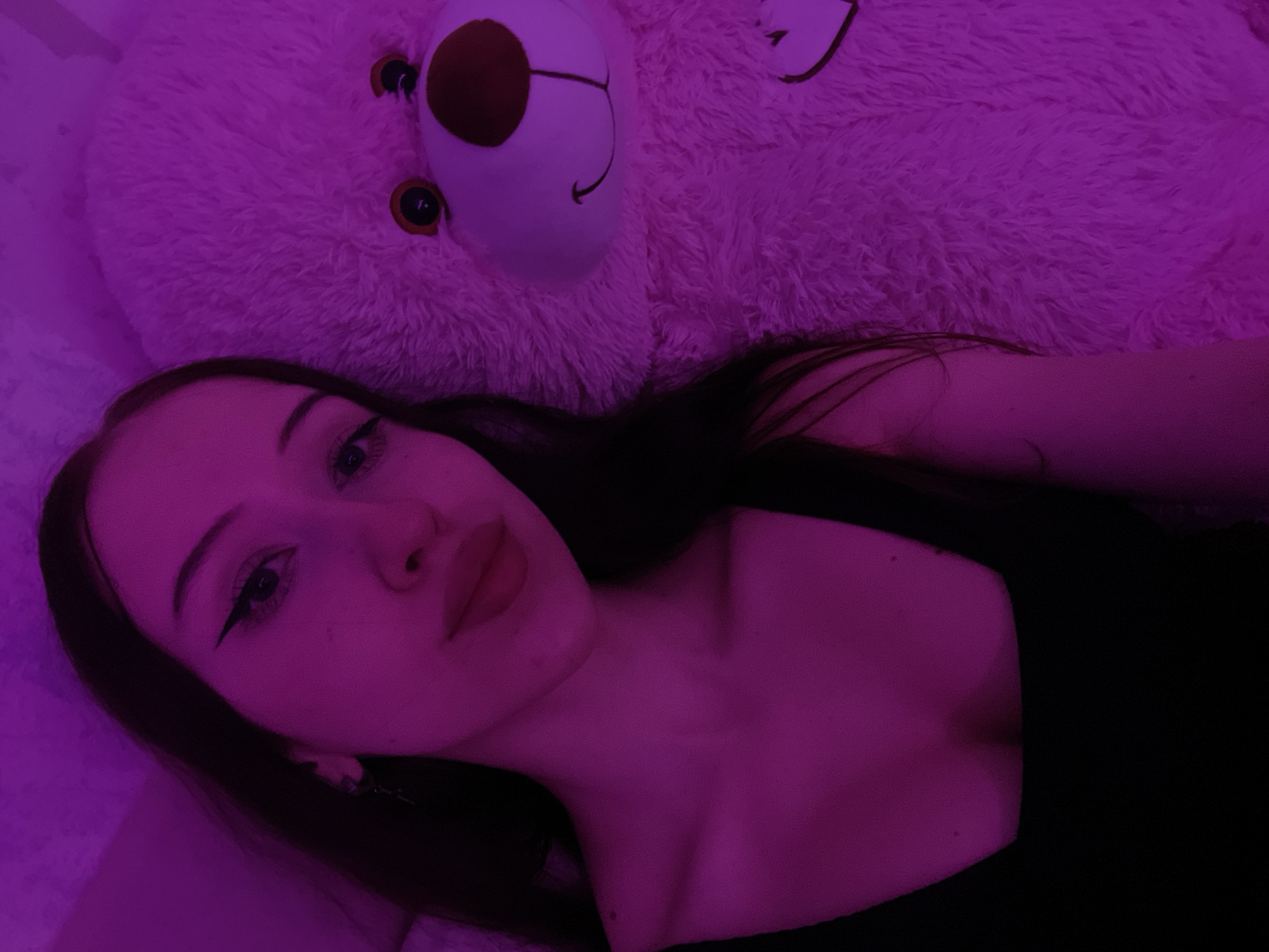 The image shows a young woman with long dark hair and makeup, including eyeliner, posing next to a large fluffy white teddy bear. The scene is illuminated by pink lighting, creating a vibrant and atmospheric effect. The woman is wearing a black top. The teddy bear has visible eyes, a nose, and a smile. There is a heart design on the teddy bear's chest. The background appears to be a wall with a slight texture. The image is a close-up shot, with the woman and the teddy bear filling most of the frame. The location is not specified in the image.
