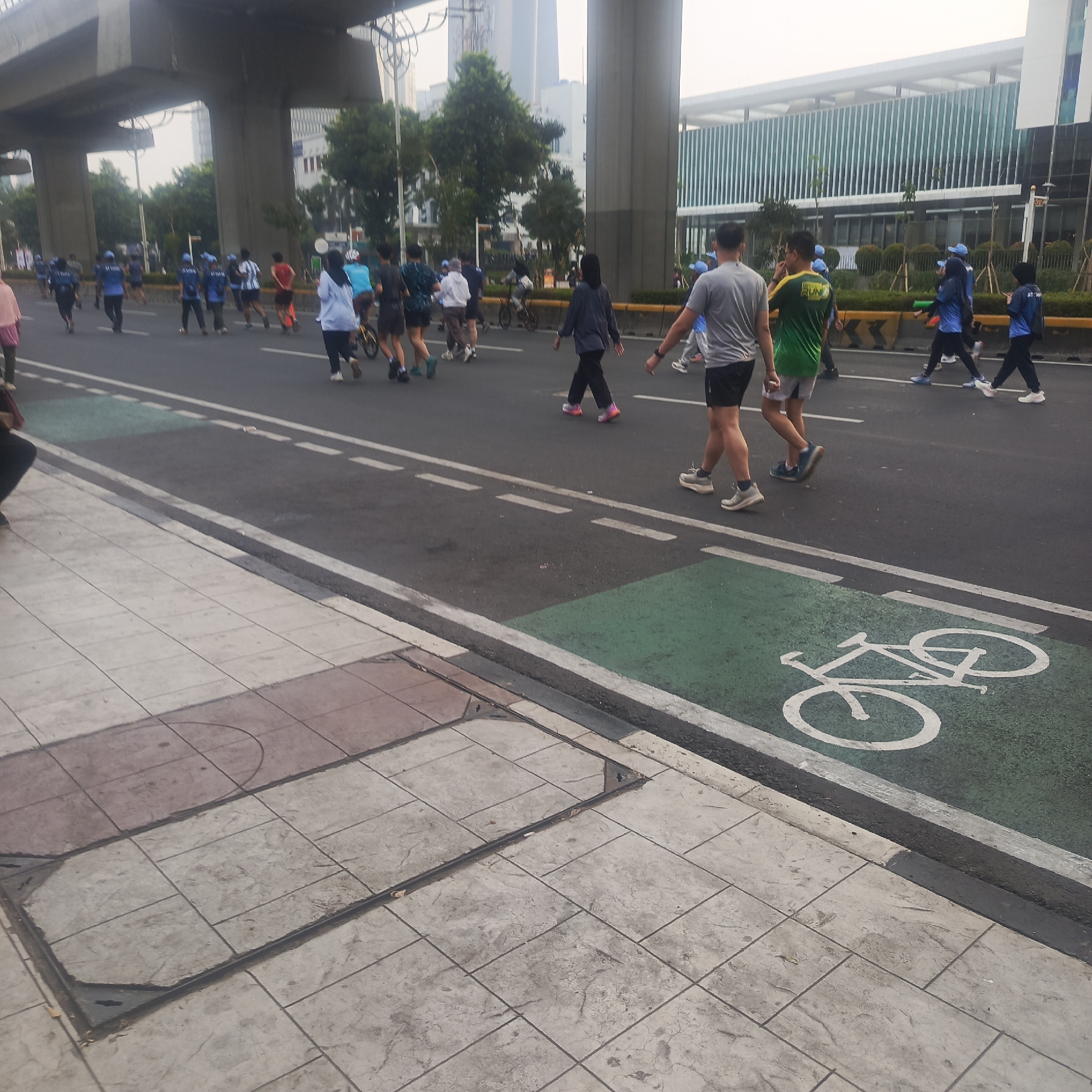 In South Jakarta, Indonesia, a group of people are walking and running on a multi-lane road with a designated bicycle lane. The road is bordered by a concrete sidewalk with patterned tiles. Overhead, a large concrete overpass structure is visible, along with modern buildings and trees in the background. The weather appears overcast. The people are dressed in athletic wear, with some wearing blue shirts and caps, indicating a possible organized event or fitness activity. One person is on a bicycle in the background, and a bicycle symbol is painted on the green bicycle lane. The scene depicts a public space used for recreation and transportation.
