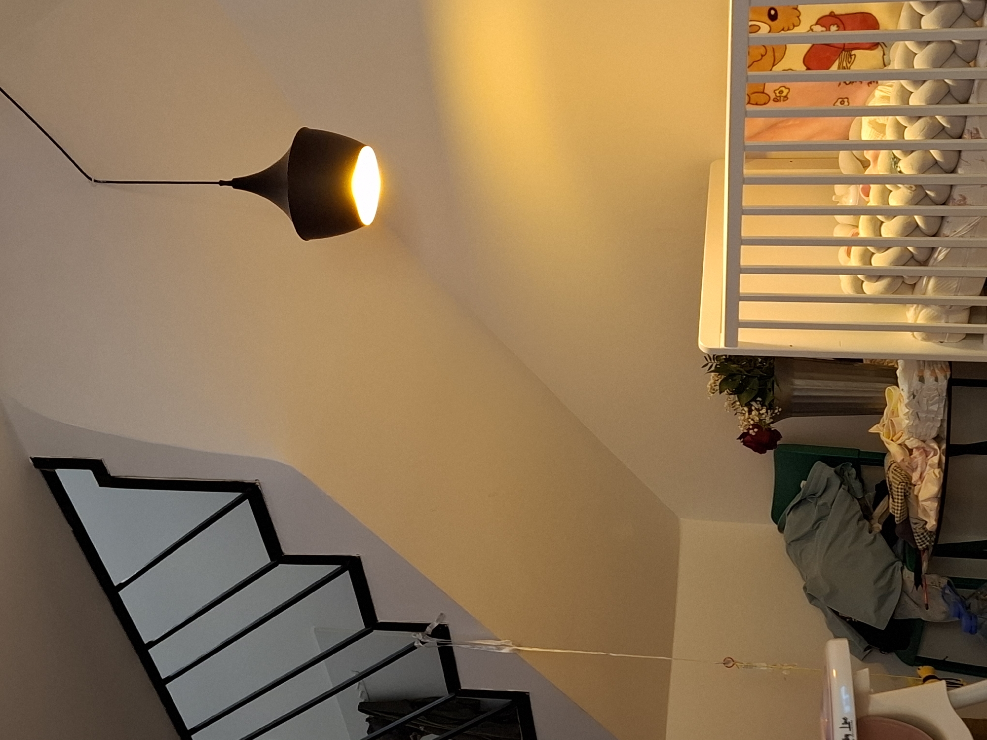 The image depicts an indoor scene within a residence in Zemun Urban Municipality, Serbia, illuminated by warm artificial light. In the upper left quadrant, a black metal staircase railing with vertical balusters ascends diagonally. Adjacent to the railing, a white wall transitions into a sloped ceiling. A thin, light-colored string with small attachments hangs vertically near the staircase on the left side.
In the upper-right area, a black, bell-shaped pendant light fixture hangs from the ceiling, emitting a bright, warm yellow light downwards, casting a glow on the right wall. The fixture's black electrical cable is visible running along the ceiling towards the upper right corner.
The lower left portion of the image shows a partial view of a white high chair or feeder. To its right, a green chair is heavily laden with folded or casually placed clothing and textiles. Adjacent to the chair, a small table with a dark frame and what appears to be a glass top holds more folded fabrics and a white container. Behind these items, a corner of the room with white walls is visible.
In the mid-right, a tall, silver-colored cylindrical vase or container holds a floral arrangement featuring a prominent red rose, other lighter-colored flowers, and green foliage.
The lower right quadrant of the image is dominated by a white wooden baby crib with vertical slats. Inside the crib, a light-colored braided bumper or guard is present along the perimeter. A baby bottle is visible among the bedding. Draped over the side or inside the crib is a colorful blanket with cartoon-like figures, including an orange cat and a red character. No individuals or animals are present in the scene.