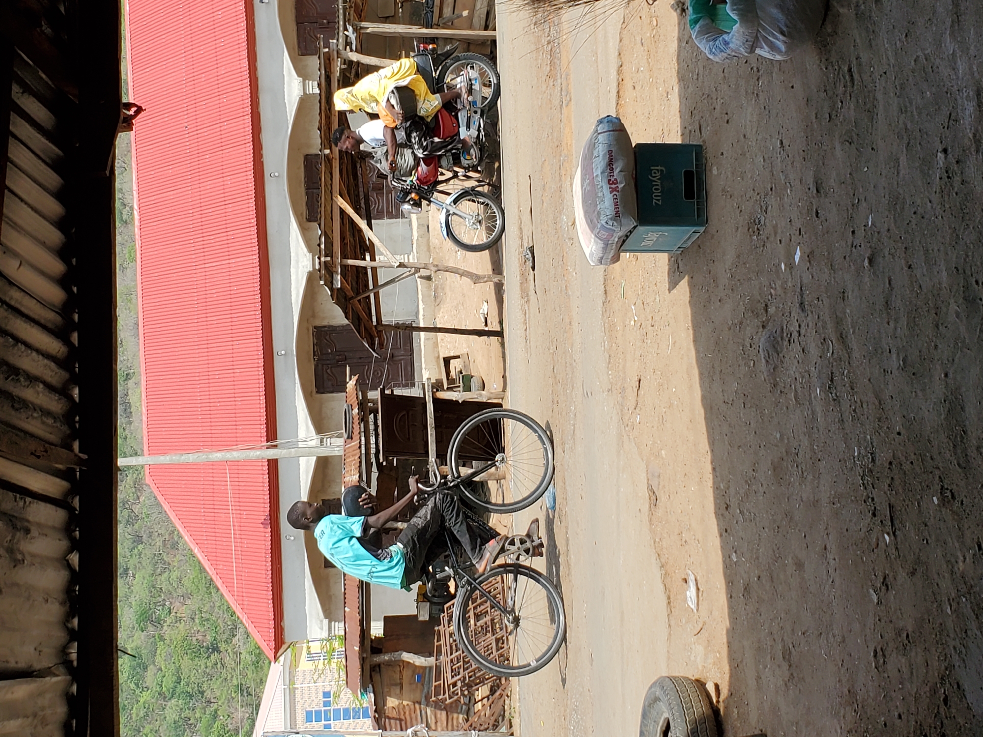 The image captures an outdoor scene in Mararaba, Nigeria, under bright daylight. The ground is composed of dry dirt and sand, with strong shadows indicating direct sunlight.
In the foreground, a person in a light blue shirt and dark pants is seated on a black bicycle, looking ahead. Further back, two individuals are near a red and black motorcycle; one wears a white shirt, and the other a yellow garment, both positioned under a makeshift wooden structure. To the right of the motorcycle, a white sack, partially labeled "DANGOTE CEMENT 3X", rests on the ground next to two stacked green plastic crates, clearly marked "Fayrouz". A lone car tire is visible on the far right.
The background is dominated by a multi-story building featuring a prominent red corrugated metal roof. The building's light-colored facade includes windows with dark wooden shutters. Beyond the settlement and building, a lush, green, tree-covered hillside stretches into the distance. The visible text includes "DANGOTE CEMENT 3X" on the sack, "Fayrouz" on the crates, and partially "VER" on the cyclist's shirt. The scene depicts a common street or compound area, suggesting daily life in the region.