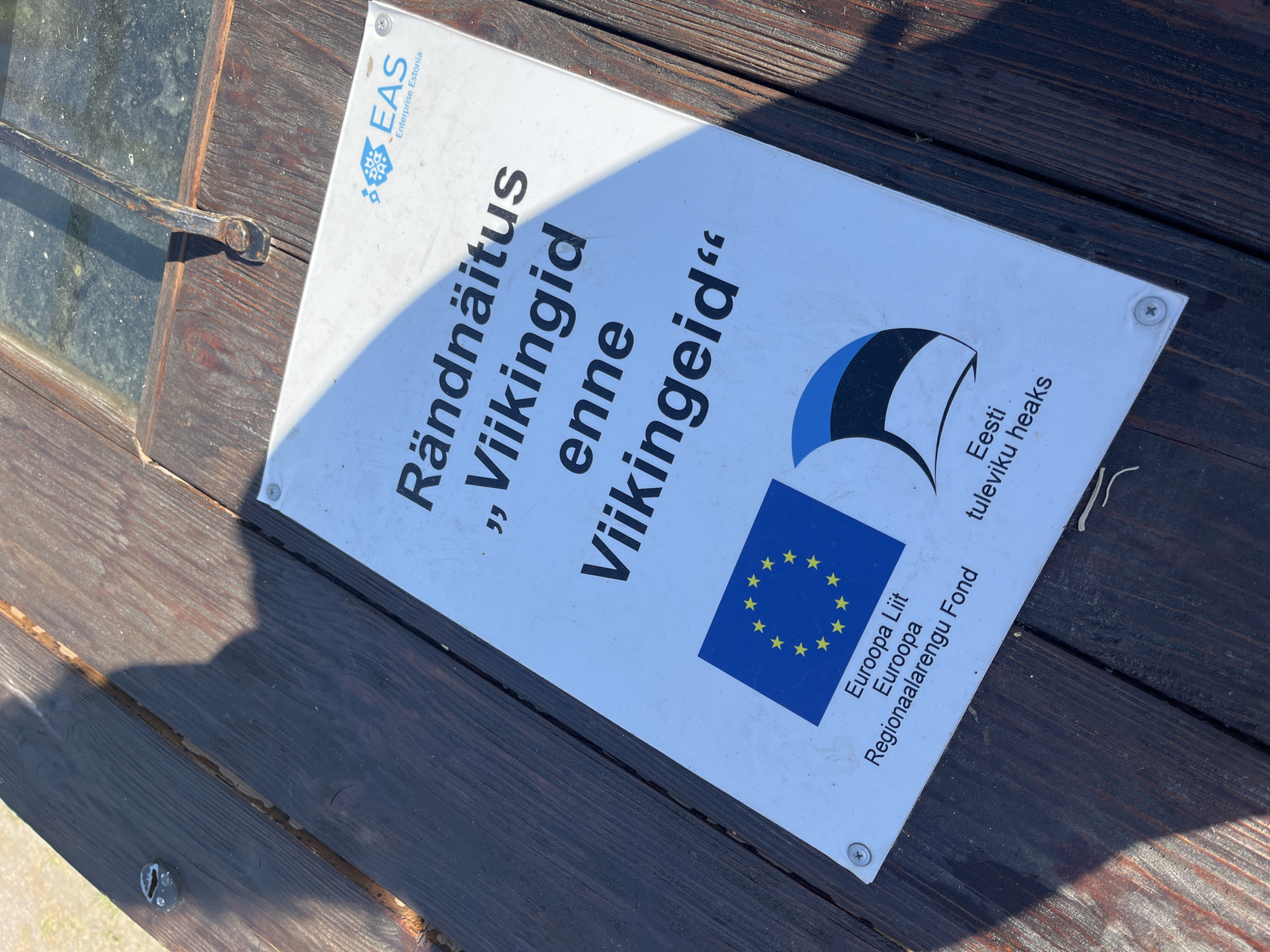 A white sign with black text and logos is affixed to a dark wooden surface. The text on the sign reads, "Rändnäitus „Viikingid enne Viikingeid" and features the logos of Enterprise Estonia (EAS), the European Union flag, and the Estonian flag. Below the European Union flag, it says "Euroopa Liit Euroopa Regionaalarengu Fond". Below the Estonian flag, it says "Eesti tuleviku heaks". The sign is angled and partially shadowed by a person, whose shadow is visible on the wooden surface. The wooden surface appears to be part of a table or bench, with visible wood grain and gaps between planks. A metal latch is visible on the wooden surface near the top right corner of the image, suggesting it might be part of a window or door. The overall scene is outdoors, with hints of sunlight and a textured background that could be dirt or gravel. The sign appears to be located in Kuressaare, Estonia, likely at an outdoor exhibition or historical site related to Vikings, given the text on the sign.