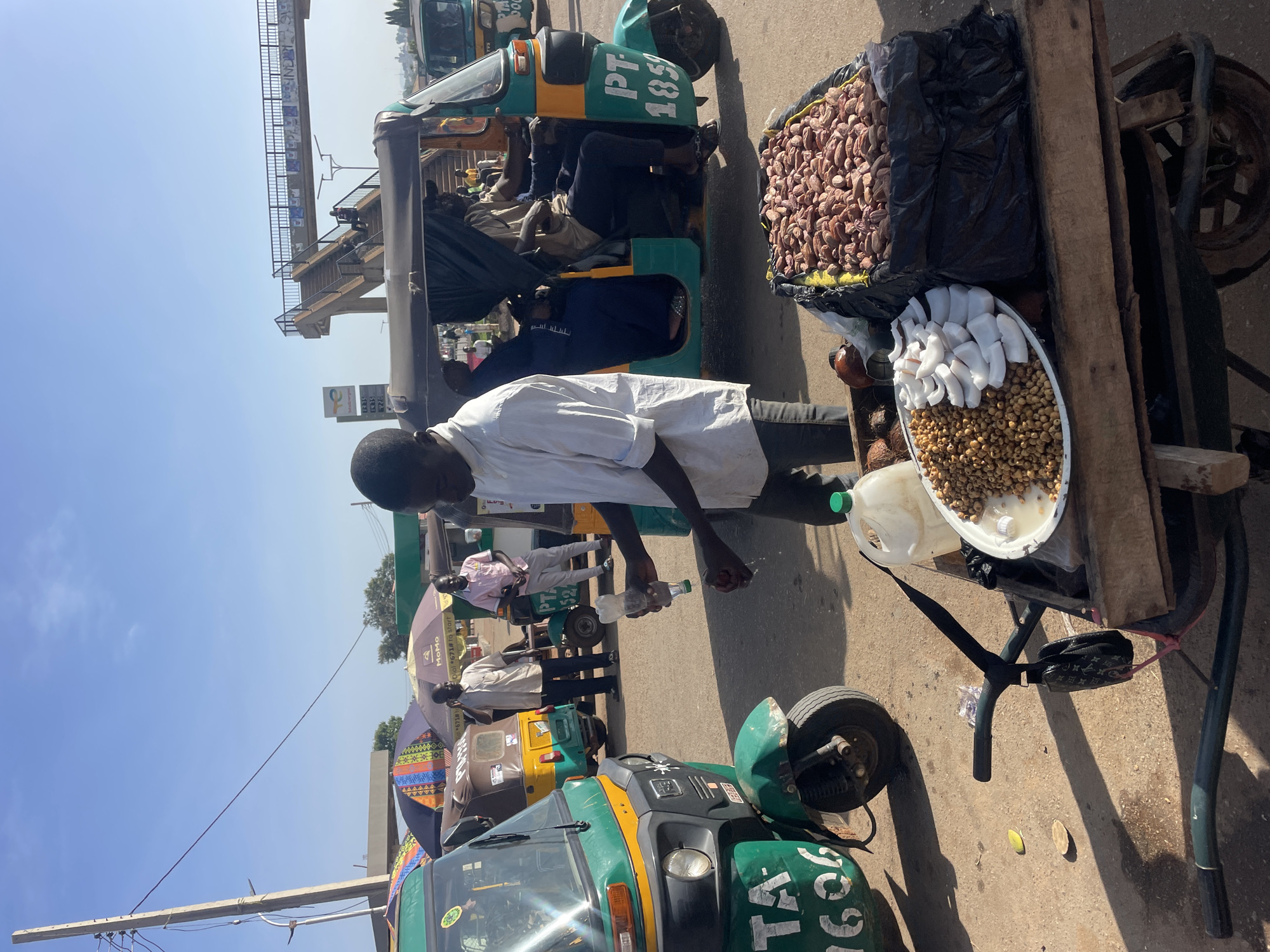 This media file captures a bustling street scene in Gura Top, Nigeria, under a bright, clear daytime sky, indicated by strong shadows. The setting is an active roadside or market area, filled with commerce and local transportation.
The central subject is a man in a white collared shirt and dark trousers, standing beside a wooden vendor cart. He is holding a clear plastic bottle, suggesting he might be selling beverages. The vendor cart is laden with produce: a large pile of light brown, oval-shaped items, possibly nuts or seeds, held within a black plastic bag. Below this, a white tray displays white chunks, resembling coconut, alongside a smaller mound of round, light brown edibles, likely groundnuts or peanuts. A white plastic jerrycan is also present on the cart.
In the background, several green and yellow/green auto-rickshaws, commonly referred to as tuk-tuks, are visible. One tuk-tuk prominently displays "TA 1606" on its side, while another further back shows "PT 1859". People are seen seated within some of these vehicles or standing nearby. Overhead, a concrete pedestrian bridge or flyover stretches across the scene, indicating urban infrastructure. Below the bridge, a "TotalEnergies" gas station sign is partially visible, displaying numbers such as "52", "1315", "1415", and "20". Several colorful umbrellas provide shade, one bearing the text "MoMo Dial *671# to start". A utility pole with wires crosses the upper left corner of the frame. The image portrays a lively scene of daily transactions and movement.