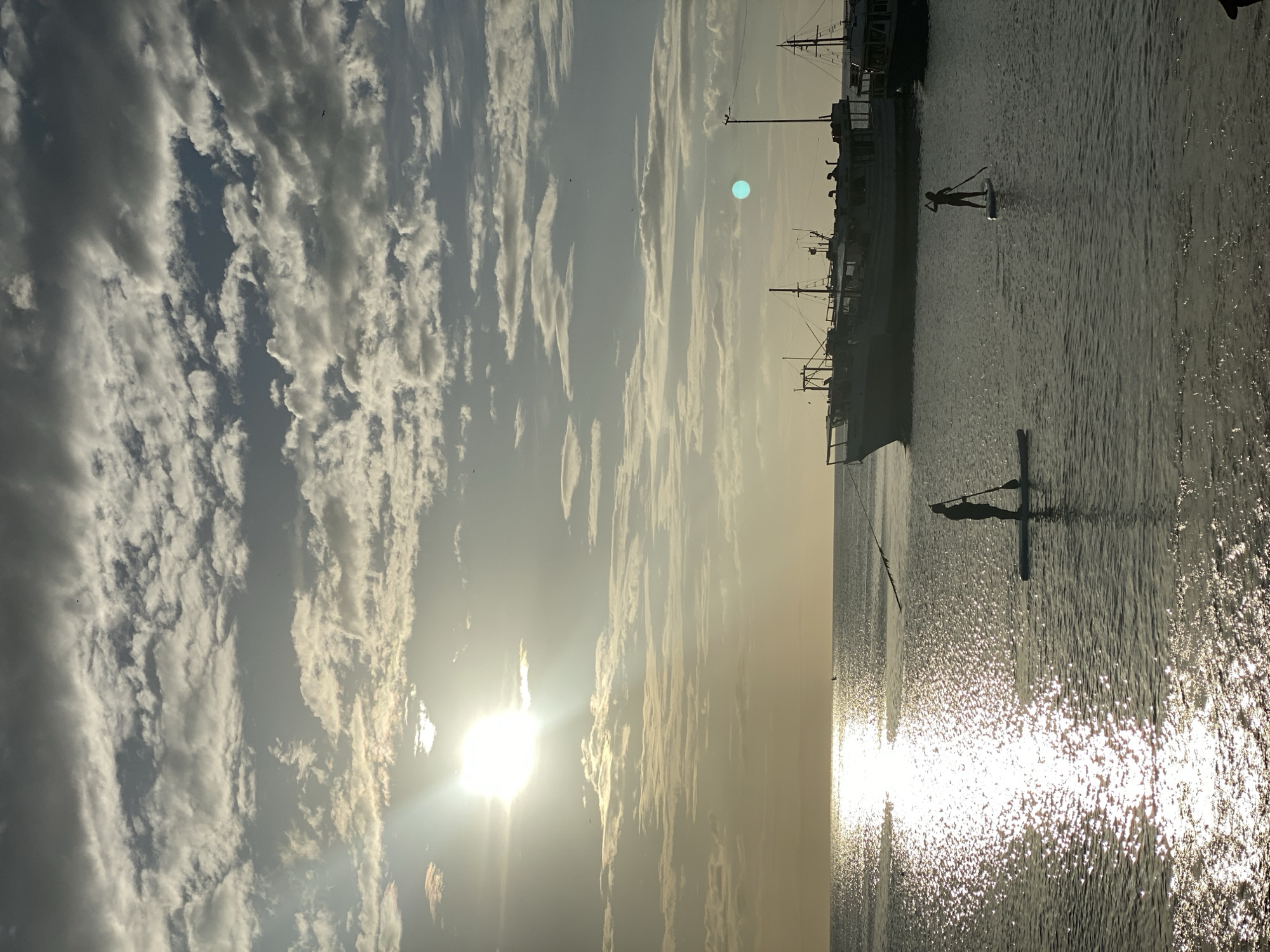 The image depicts a sunset over the water in Juan Griego, Venezuela. Two individuals are stand-up paddleboarding in the foreground, silhouetted against the bright, shimmering water. In the mid-ground, several boats are anchored, their forms dark against the horizon. The sky is filled with a dramatic display of clouds, illuminated by the setting sun. The sun itself is a brilliant orb, casting a strong glare on the water.