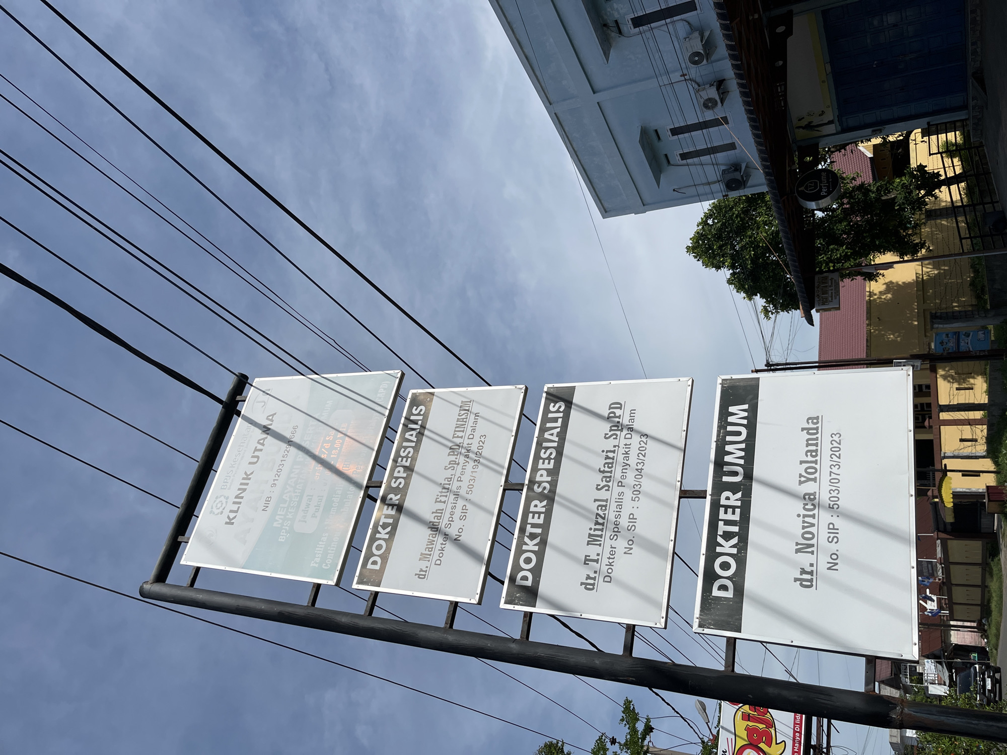 The image shows a pole with multiple signs in front of a light blue building under a cloudy sky. The signs are for "KLINIK UTAMA," which is a primary clinic. There are also signs indicating "DOKTER SPESIALIS" (Specialist Doctor) and "DOKTER UMUM" (General Practitioner). One specialist doctor listed is dr. Mawaddah Fitria, Sp.PD, FINASIM, specializing in internal medicine, and another is dr. T. Mirzal Safari, Sp.PD, also specializing in internal medicine. A general practitioner listed is dr. Novica Yolanda. The signs also include National Identification Numbers (NIB) and SIP (Surat Izin Praktik - Practice Permit) numbers with dates. In the background, there are other buildings, trees, and a street with a car. The environment appears to be a commercial or residential area. The overall scene is a street view of a clinic with its service information displayed. This is likely a street in Lhokseumawe, Indonesia, given the context of the clinic's name and the typical signage seen in Indonesian cities.