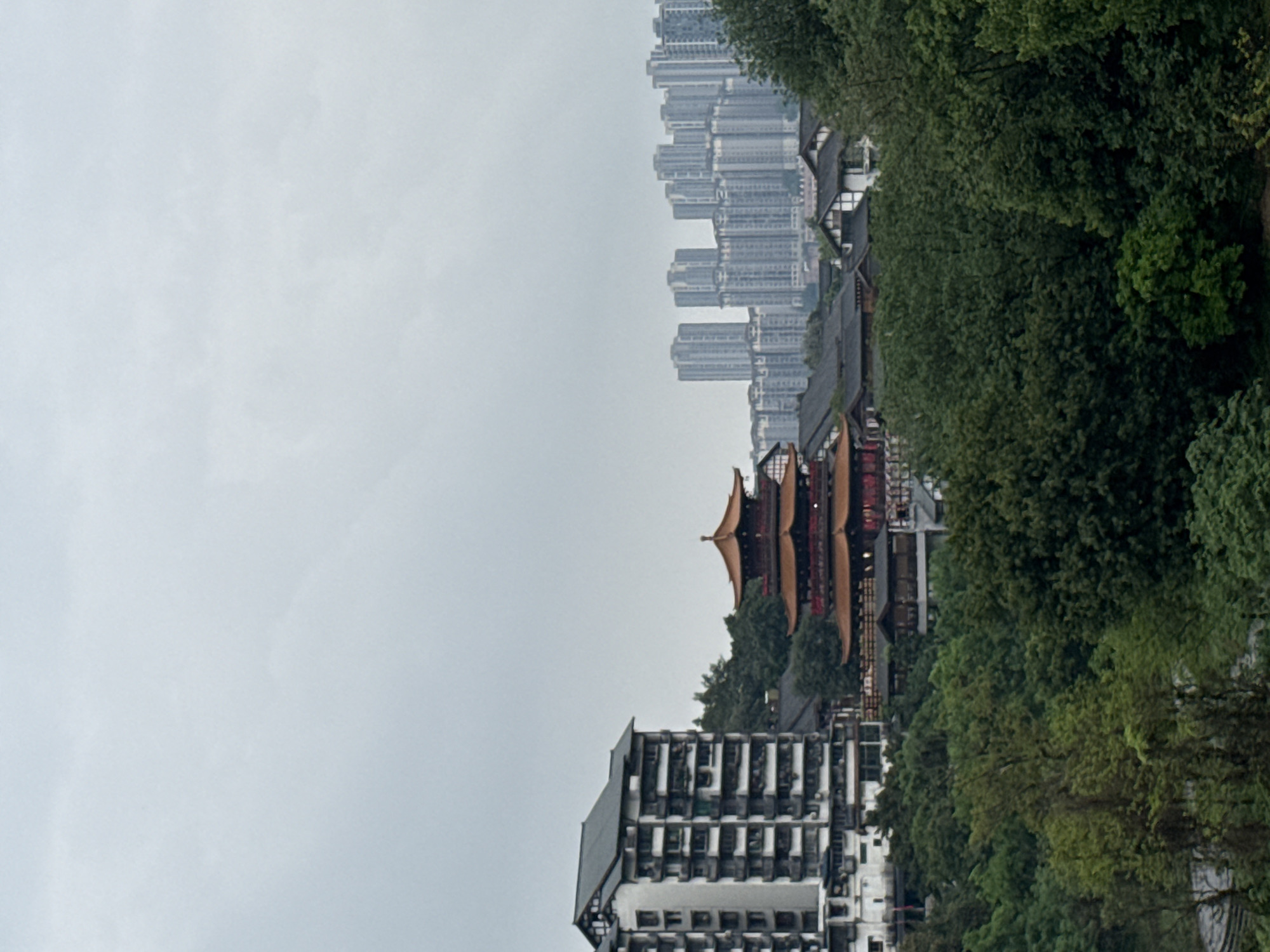 The image depicts a view of Shapingba District, China, showcasing a mix of modern and traditional architecture. High-rise apartment buildings line the horizon under an overcast sky. A traditional-style Chinese pagoda stands out amidst the buildings and surrounding lush greenery. A modern multi-story building is also visible.