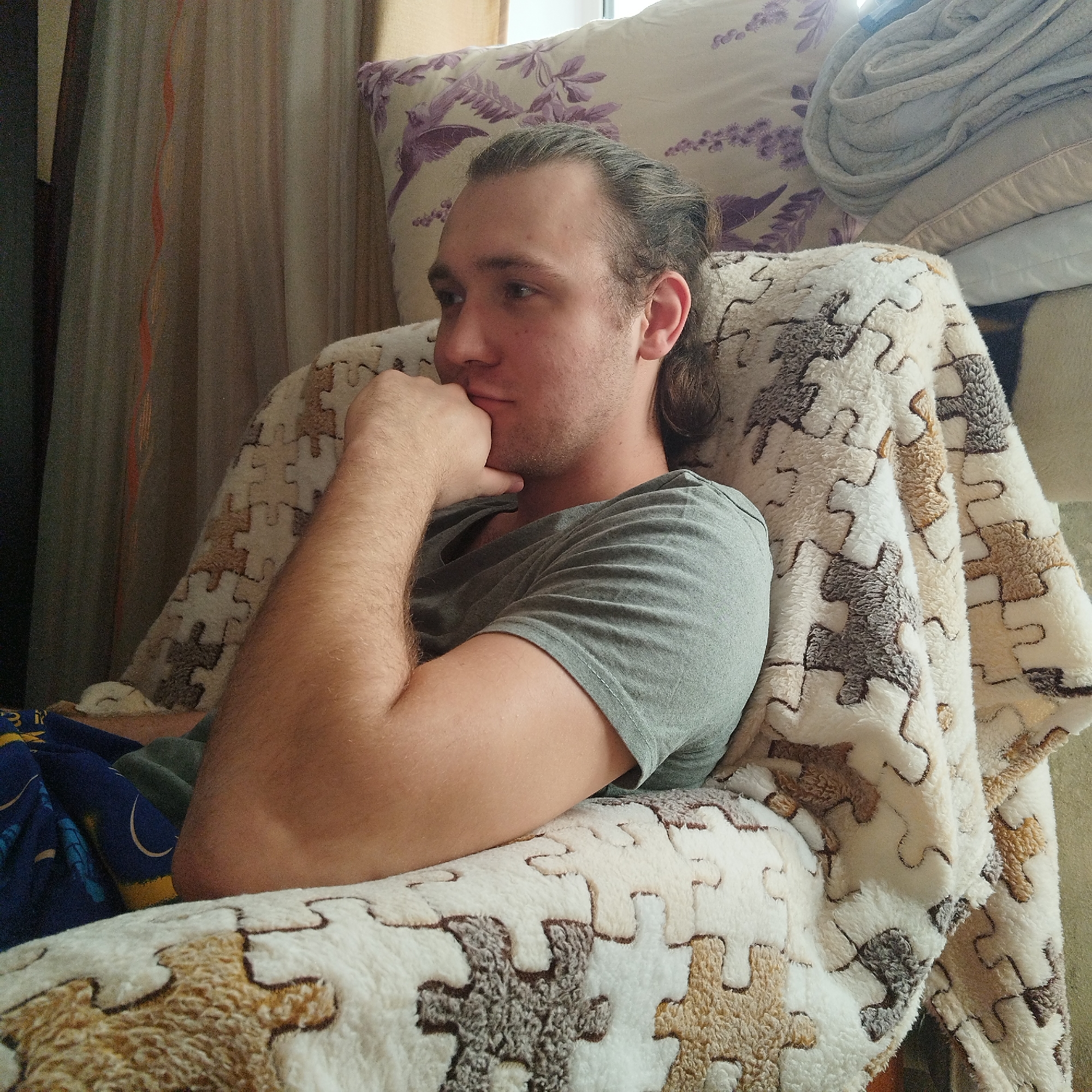 A light-skinned man with brown hair pulled back into a low ponytail sits in an armchair covered with a puzzle-piece patterned blanket. He wears a gray t-shirt and rests his chin on his hand, looking to the left. Behind the chair are pillows, one with a floral pattern, and a stack of folded towels or blankets. Curtains are visible in the background near a window. The location is in Yekaterinburg, Russia.