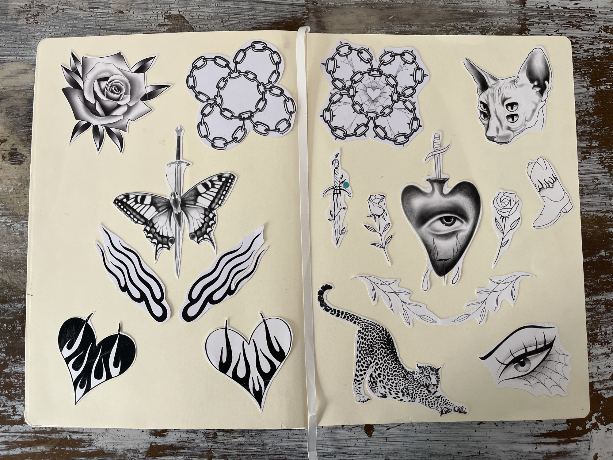 A two-page spread in a cream-colored sketchbook displays a collection of black-and-white, tattoo-style drawings or stickers.
On the left page, from top to bottom, there is a detailed rose, a chain design, a butterfly impaled with a sword, two flame-detailed shapes, and a heart.
On the right page, from top to bottom, there is another chain design, a four-eyed cat, a sword over a heart, a rose, a boot, a leopard, and an eye.
The sketchbook rests on a weathered wooden surface. The images look to be a collection of tattoo designs from the 1990's.
There is no indication of any activity or environment to suggest where this was taken place in Barcelona, Spain. It is likely that the location is not Barcelona, Spain.