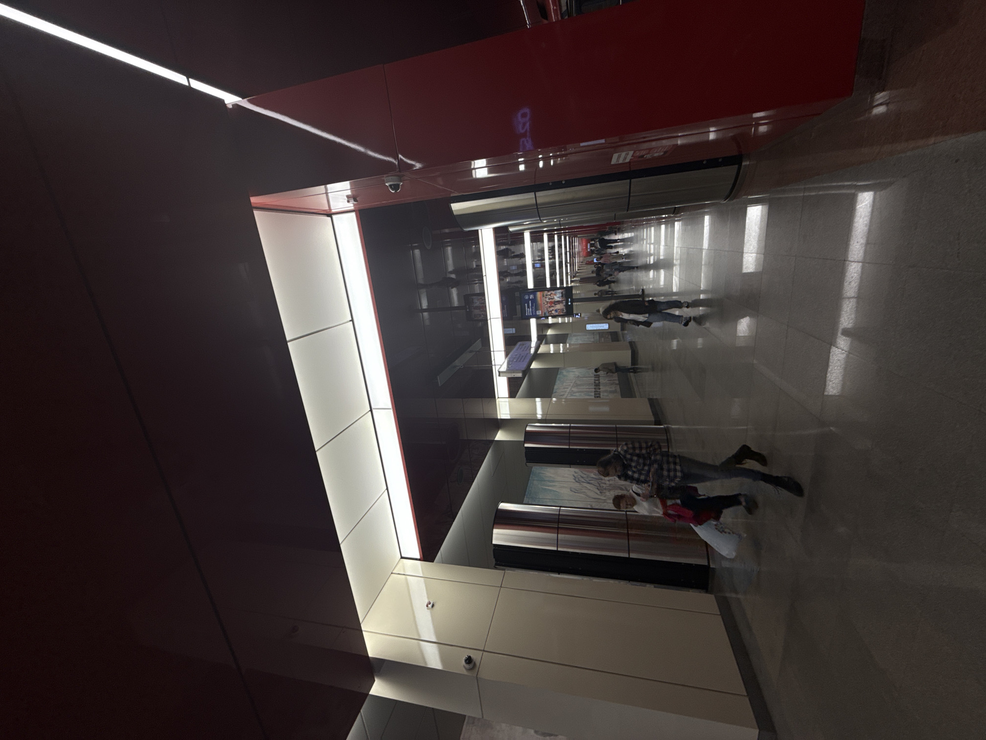 The image depicts a contemporary metro station in Moscow, Russia, characterized by its modern design. The ceiling is a deep red, with linear white light fixtures providing illumination. The walls are a mix of neutral tones and polished metallic surfaces, with large, abstract murals adorning some sections. The floor is highly reflective, mirroring the overhead lights and the architecture of the station.
In the foreground, two individuals are walking away from the camera. The person on the left is wearing a dark top and lighter pants, carrying a white plastic bag. The person on the right, dressed in a plaid shirt and dark pants, appears to be looking at their phone.
Further down the platform, several other individuals are visible, walking in various directions. The overall scene conveys a sense of urban transit and daily life in the Moscow metro system. Security cameras are mounted on some of the vertical structural elements.