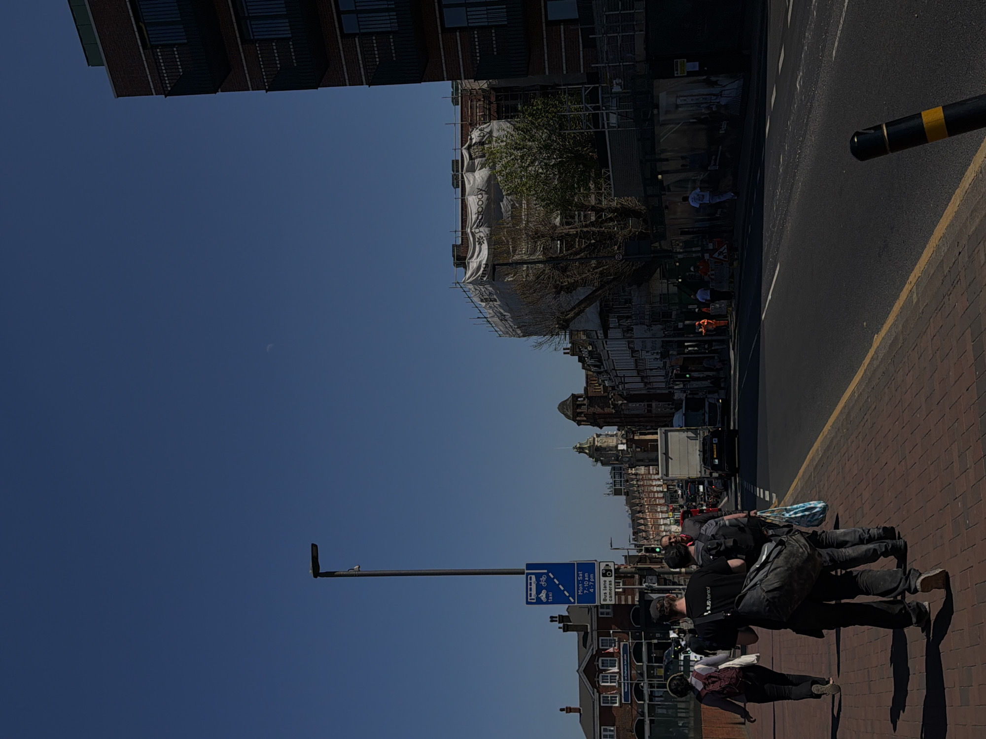 A daytime, low-angle, rotated view of a street scene in what appears to be London, United Kingdom. The scene is oriented sideways. The sky is a clear blue and takes up more than half of the left side of the image. Along the bottom of the image is a pedestrian brick walkway with a few pedestrians present. Three individuals are walking, they are generally wearing casual clothes.
A two-lane street runs parallel to the walkway, with a solid white line dividing the two lanes and a yellow line marking the edge of the roadway. A black and yellow bollard is present in the upper right corner. Vehicles are traveling on the road; a large white truck is visible in the center of the image.
On the far side of the street, buildings of varying heights and architectural styles line the street. Scaffolding and white cloth are partially obscuring some of the buildings. A tall street light pole is seen on the left side. A blue sign indicating it's a bus lane with specific hours of operation is attached to the pole.