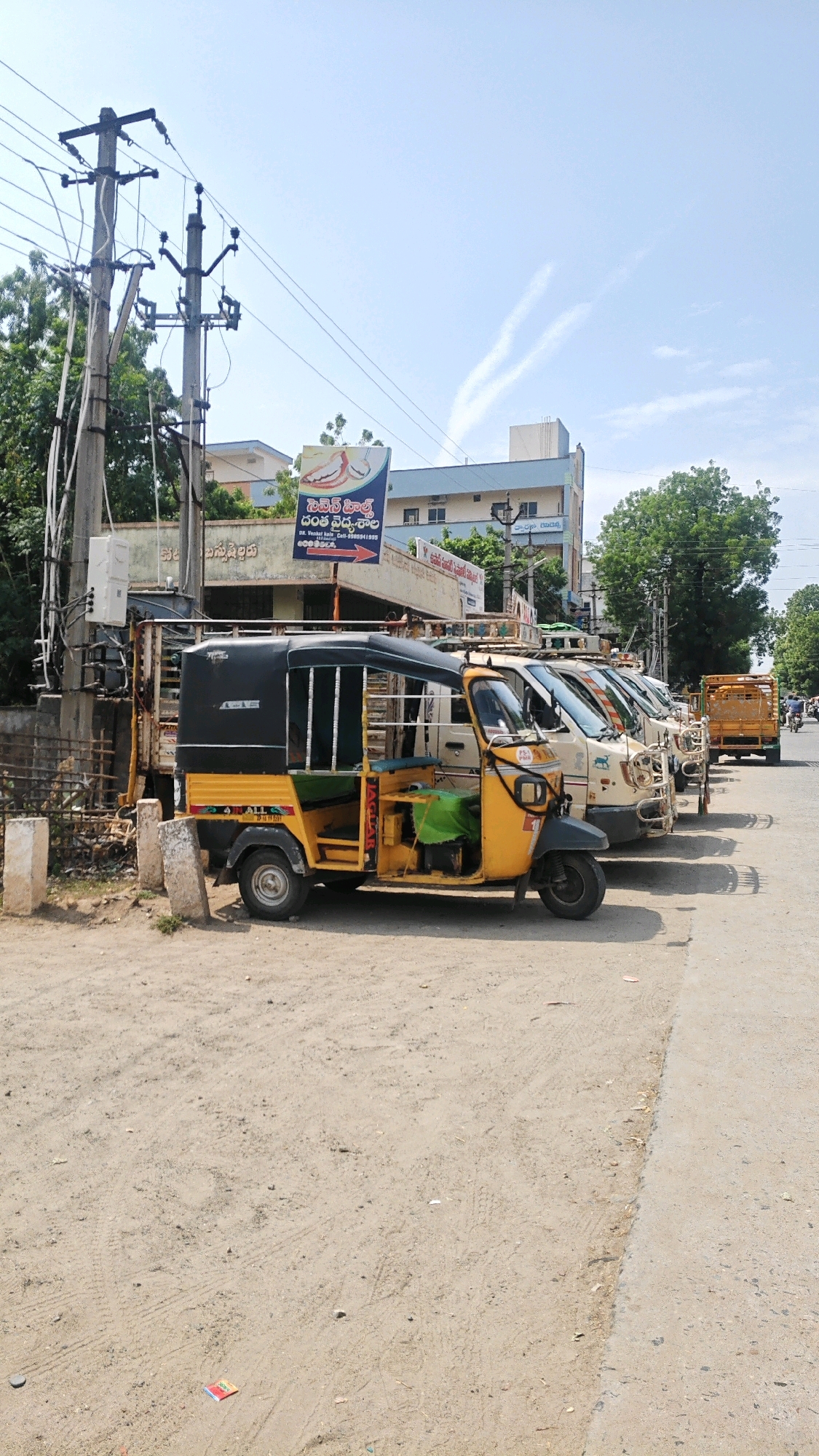 In Virat Nagar, India, a sunny daytime scene unfolds on a dusty road lined with parked vehicles. In the foreground, a yellow and black auto-rickshaw is parked, its canvas roof and side panels visible. Behind it, a row of cream-colored vans are also parked, their fronts facing towards the right side of the frame. Further down the road, a yellow truck with an open cargo bed is also parked. Electrical poles and wires dominate the left side of the image, with a blue and white building visible in the background. Trees and foliage add greenery to the environment. The road itself is a mixture of dirt and a paved section on the right. The sky is a clear blue with faint white contrails from an aircraft.