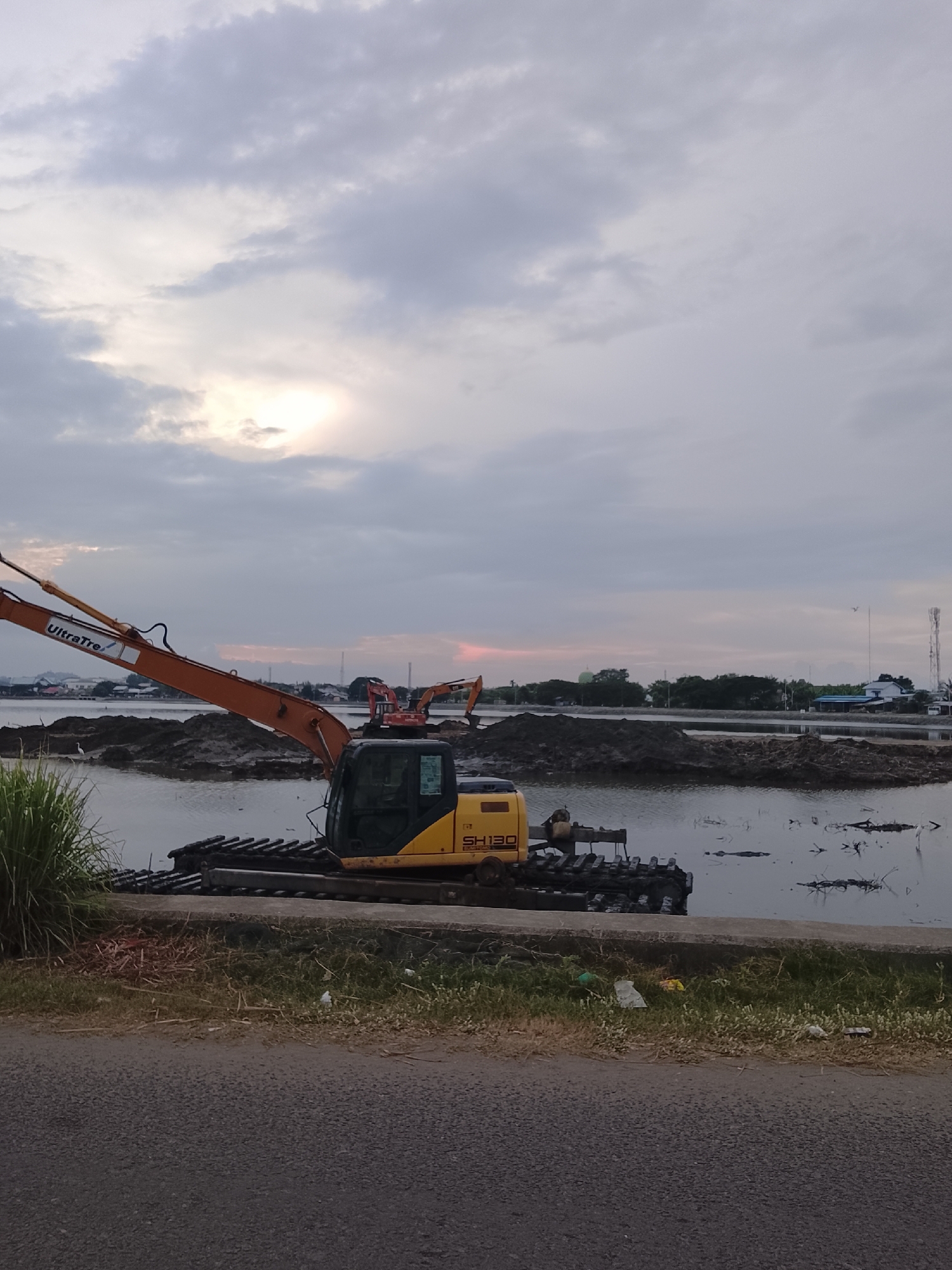 The image shows a construction site near the coast in Lhokseumawe, Indonesia. A yellow excavator, identified by the "UltraTre" logo and "SH130" model name, is partially submerged in water, positioned on a muddy bank. The excavator's arm is extended, and it appears to be actively working or recently used. In the background, other construction equipment and piles of dirt are visible, suggesting ongoing land reclamation or infrastructure development. The sky is overcast with the sun peeking through the clouds, casting a soft light on the scene. The environment appears to be a coastal area with some vegetation and distant buildings and industrial structures, hinting at the proximity to the city's port or industrial zones. The overall impression is one of a working industrial landscape at dusk or dawn.