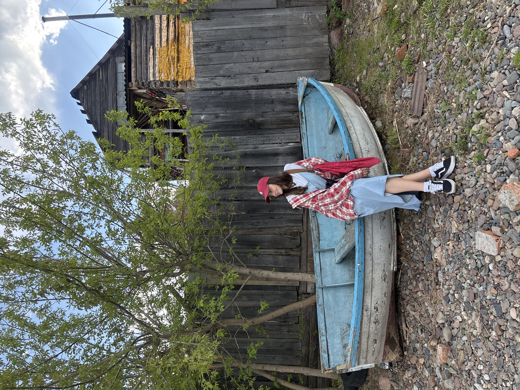A young woman wearing a red cap, a white t-shirt, a denim dress, and a red and white plaid shirt is sitting in a light-blue boat. The boat is partially nestled against a weathered, wooden building with a yellow section. A tree with green leaves stands to the left of the building and boat. The ground surrounding the boat is covered in small rocks and some patches of grass. The background reveals a blue sky with clouds. This image was taken in the city of Овсянка, Russia.