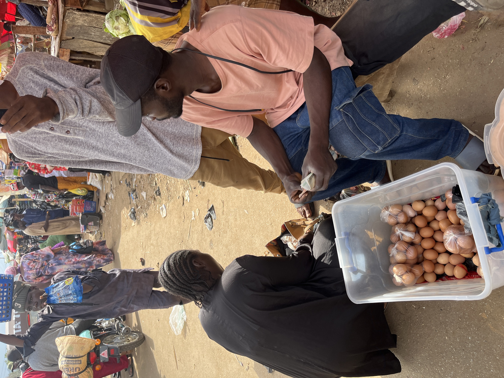 The image depicts an outdoor market scene in Rafin Par, Nigeria. In the foreground, a person in a black abaya is seated, attending to a brown leather bag. Beside them is a clear plastic container filled with numerous brown eggs, some loose and others bagged. To the right, a man wearing a light pink t-shirt, blue jeans, and a dark baseball cap is seated, holding a wad of cash. Another person, partially visible, stands behind him. In the background, the market is bustling with activity. A person in a grey robe and hat holds a blue basket and a plastic bag aloft. Several stalls with goods, including food items and motorcycles, are visible. The ground is dusty, with scattered debris. The overall environment suggests a lively and typical day at a Nigerian market.