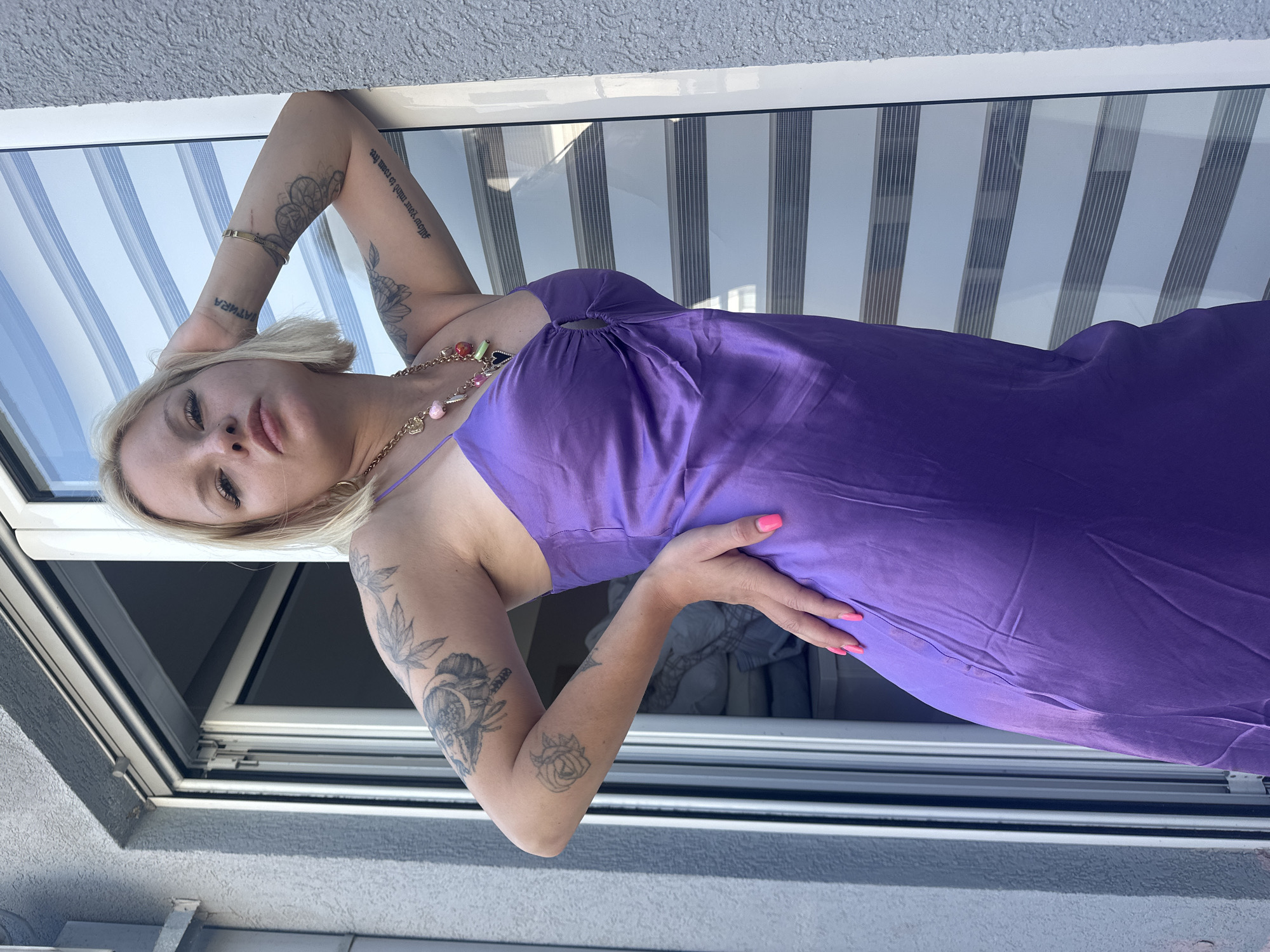 A woman with blonde hair and tattoos on her arms is posing for a photo. She is wearing a purple dress and a necklace with beads. The woman is leaning against a window frame, with a striped blind in the background. The scene appears to be outdoors, possibly on a balcony or terrace, given the presence of a window and a textured wall. The lighting suggests it is daytime, likely sunny. The woman's expression is relaxed, and she is looking towards the camera. The image is taken from a slightly elevated angle, looking down at the woman. There is visible text on the woman's arm.