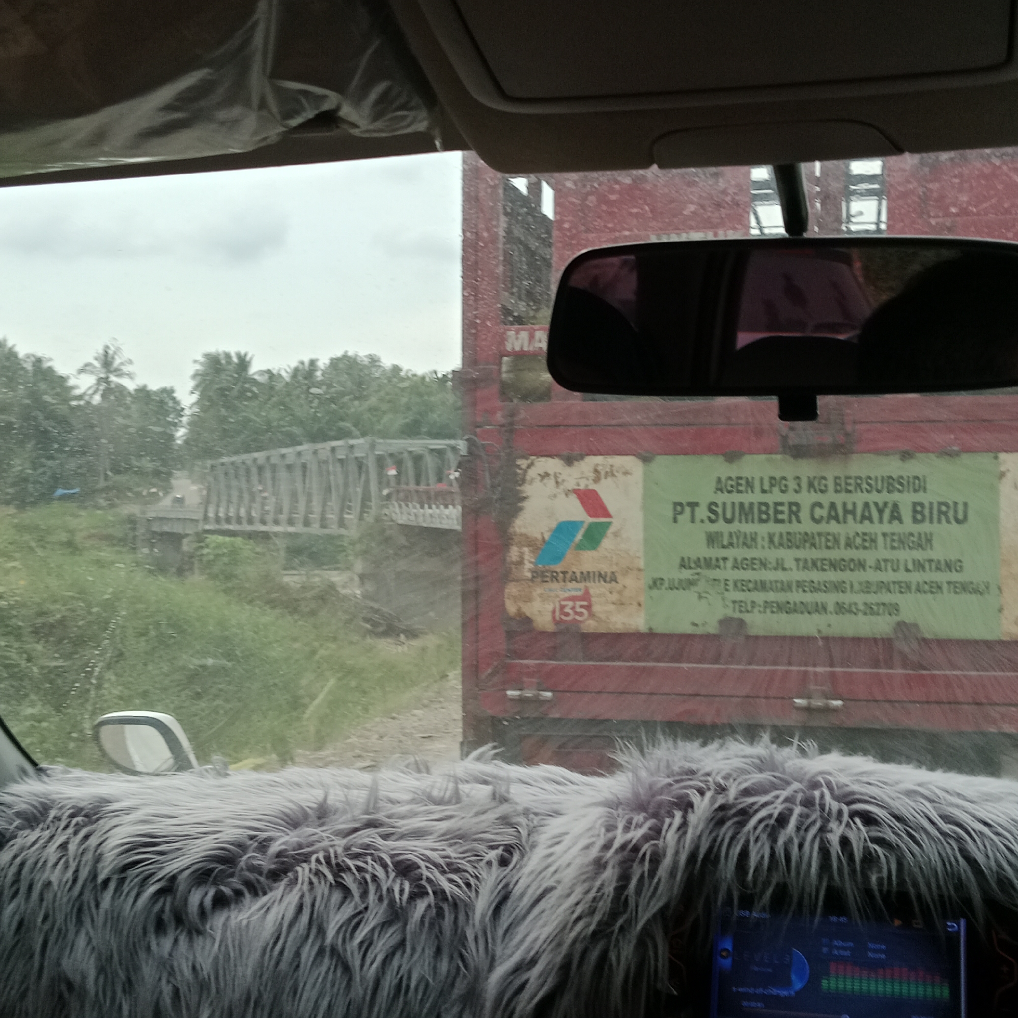 The image, taken from inside a vehicle, shows a view of a road with a bridge in the distance and a red truck on the right. The truck has a sign that says "AGEN LPG 3 KG BERSUBSIDI PT. SUMBER CAHAYA BIRU WILAYAH: KABUPATEN ACEH TENGAH". The environment appears to be rural with lush green vegetation and trees. The sky is overcast, suggesting it might be raining or the weather is generally gloomy. The foreground of the image is dominated by a fuzzy grey material, likely a car accessory, and a screen displaying what appears to be audio level meters. The bridge in the background is a truss bridge made of metal. There are no discernible people clearly visible in the scene. The overall impression is a snapshot of a journey through a somewhat remote area in Indonesia.