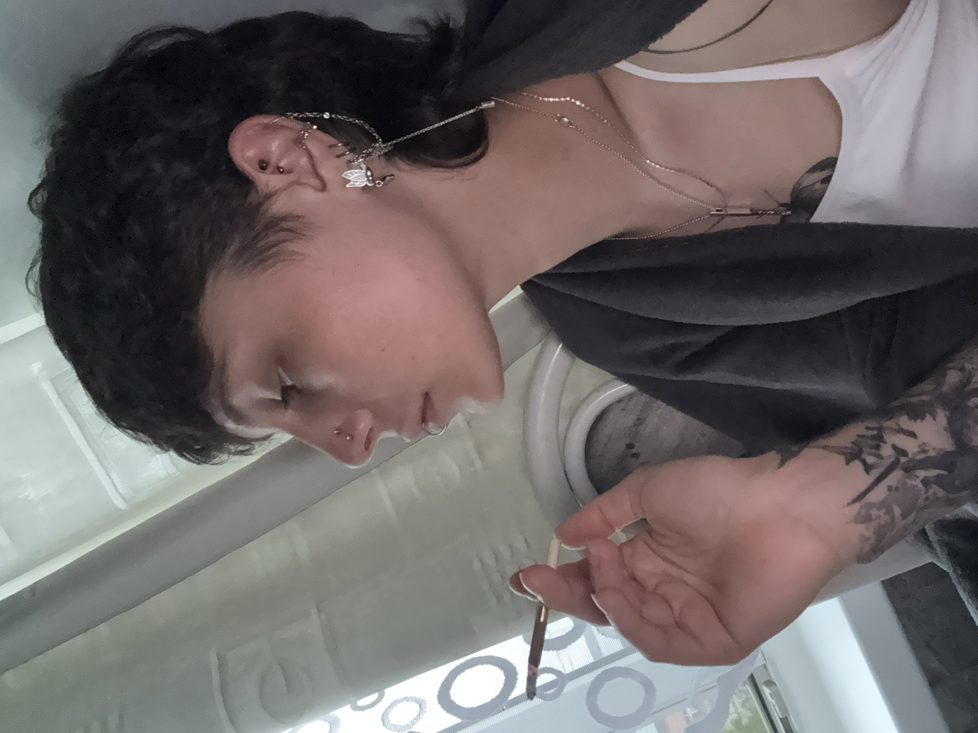 A young individual with short, dark, curly hair styled in a mullet cut is shown in side profile, indoors, in Saint Petersburg, Russia. The person holds a hand-rolled cigarette, featuring a dark wrapper and light-colored filter, between the thumb and index finger of the left hand, positioned near the mouth. The nose displays two stud piercings, and the lower lip has a hoop piercing. The visible left ear features an industrial piercing, multiple studs, and an elaborate ear cuff combining a butterfly motif near the lobe with chains extending to upper ear elements and a dangling chain. Two delicate chain necklaces, one with a small pendant, are visible. A dark, artistic tattoo covers the left forearm. The individual wears a white tank top under a dark grey or black open cardigan. The background includes a window on the left with a white frame and a blind featuring large hollow circular patterns. Natural light from the window illuminates the scene.