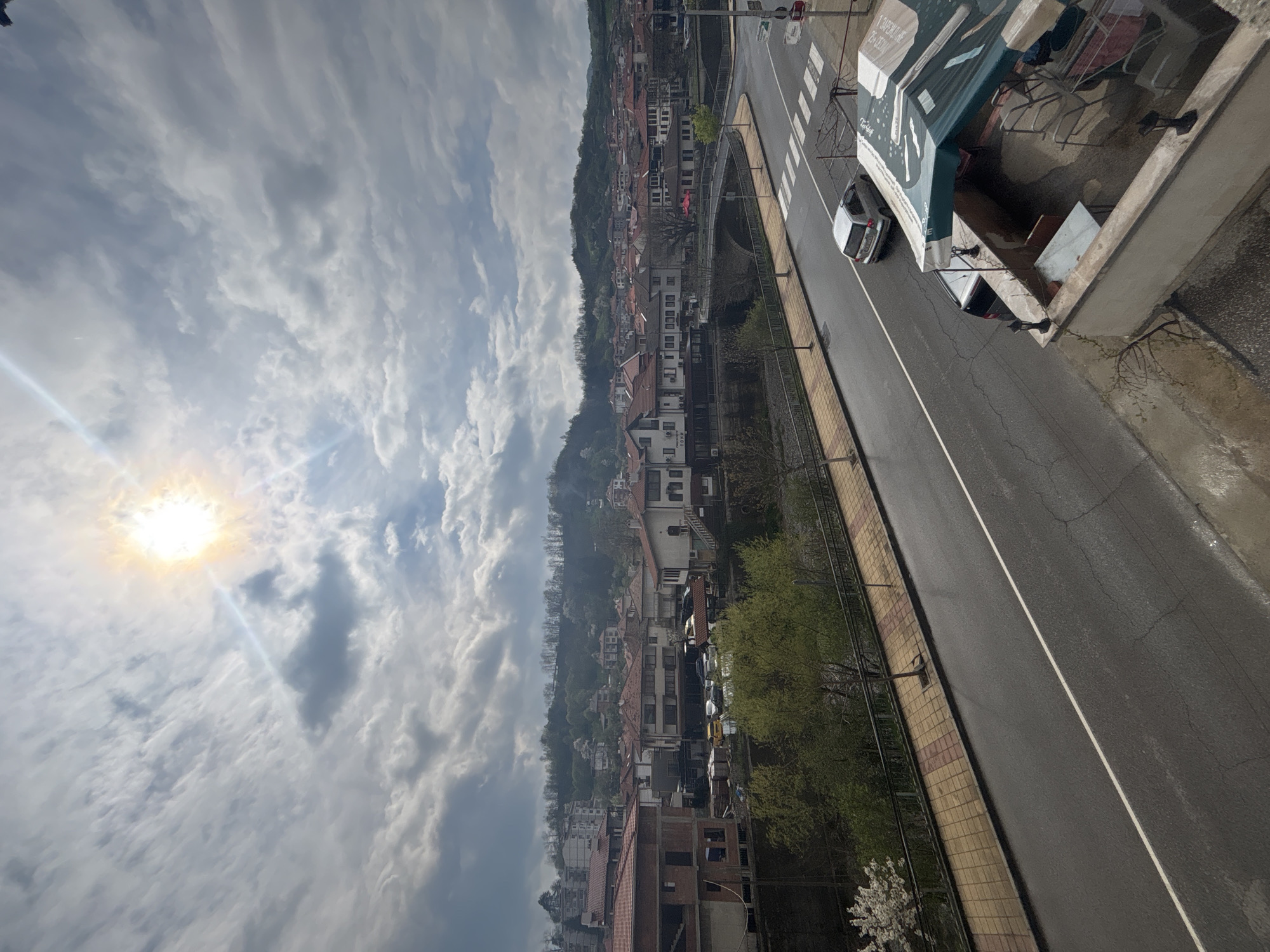 The image presents an elevated, panoramic view of a town, identified as Zlatograd, Bulgaria, under a partly cloudy sky during daytime. The sun, partially obscured by clouds, shines brightly from the upper left, creating a visible lens flare.
The sky occupies a significant portion of the upper frame, filled with a mix of streaky grey and white clouds, with patches of clear blue sky visible. Below the sky, the town sprawls across the mid-ground, featuring numerous buildings with distinctive red-tiled roofs nestled against a lush green, tree-covered hillside that extends into the distant horizon. The architecture appears characteristic of a European hillside town.
In the foreground and mid-right, an asphalt road runs diagonally, marked with a solid white line and a prominent pedestrian zebra crossing. A patterned sidewalk, made of yellow and terracotta tiles, borders the road on the left side. Several vehicles are visible: a silver car is parked near the zebra crossing, and a darker vehicle is partially seen under a green awning.
The viewpoint appears to be from a concrete balcony or terrace, the edge of which is visible in the immediate right foreground. This area includes a green-and-white striped awning. Visible text on the awning clearly reads "1 ЗАВЕДЕНИЕ" (1 Establishment/Restaurant) and a phone number "25-39-37". Underneath the awning, outdoor furniture such as chairs and a table, along with a clothes drying rack, suggest an outdoor living or commercial space.
Beyond the road, to the left, vibrant green trees and shrubs line the embankment, including a bush with delicate white blossoms. A pedestrian bridge, featuring a distinct red structural element, spans a gap between buildings further into the town. The overall scene conveys a calm, everyday atmosphere in a picturesque, sunlit hillside community.