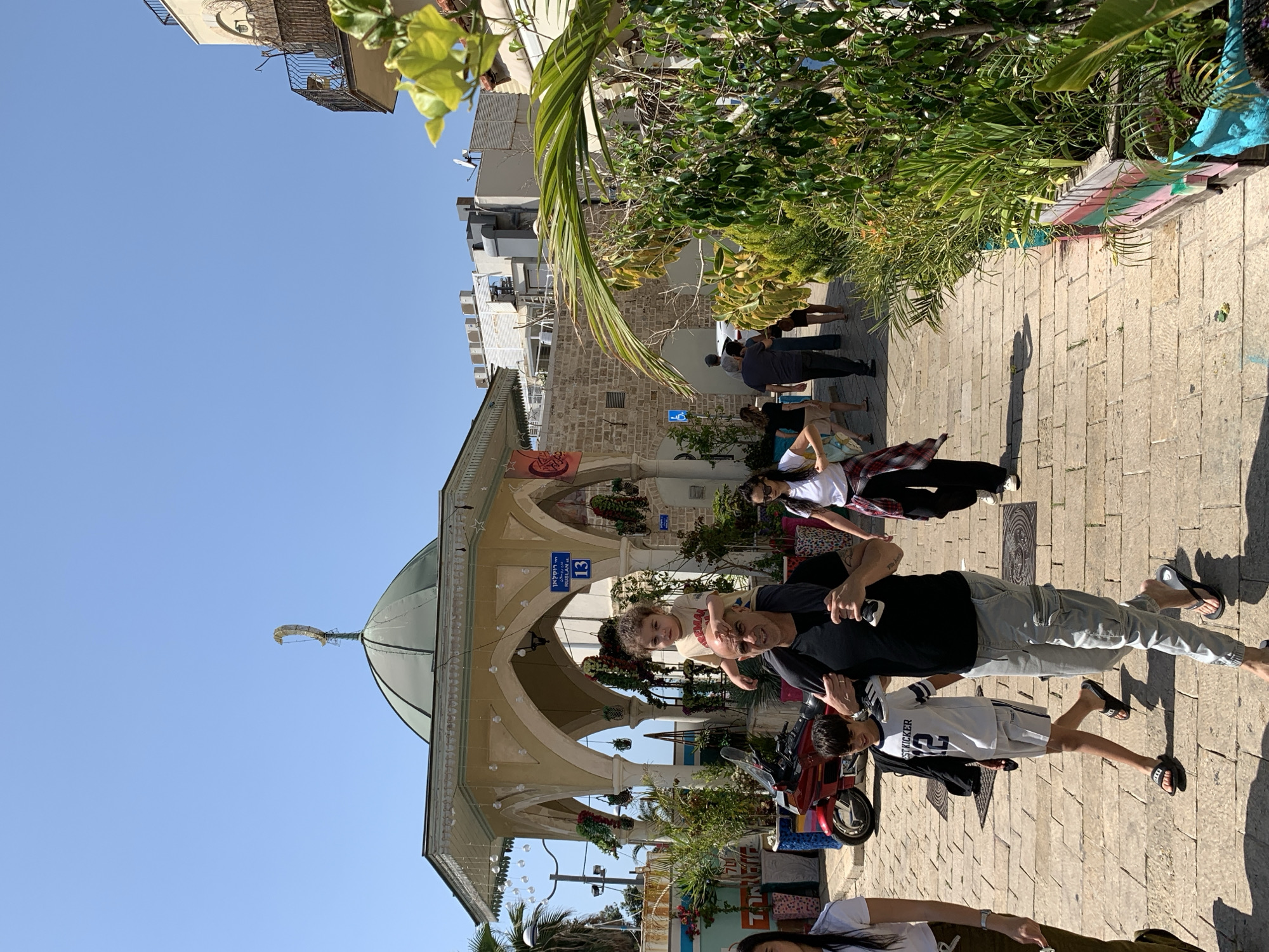 The image shows a street scene in Jaffa, Tel Aviv, Israel. The foreground features a cobblestone street with people strolling. A man is carrying a young child on his shoulders while walking down the street with a younger boy. Behind them are other pedestrians. To the left is a building with arches, decorated with greenery and strings of lights. The background includes more buildings and foliage, with a clear blue sky above.