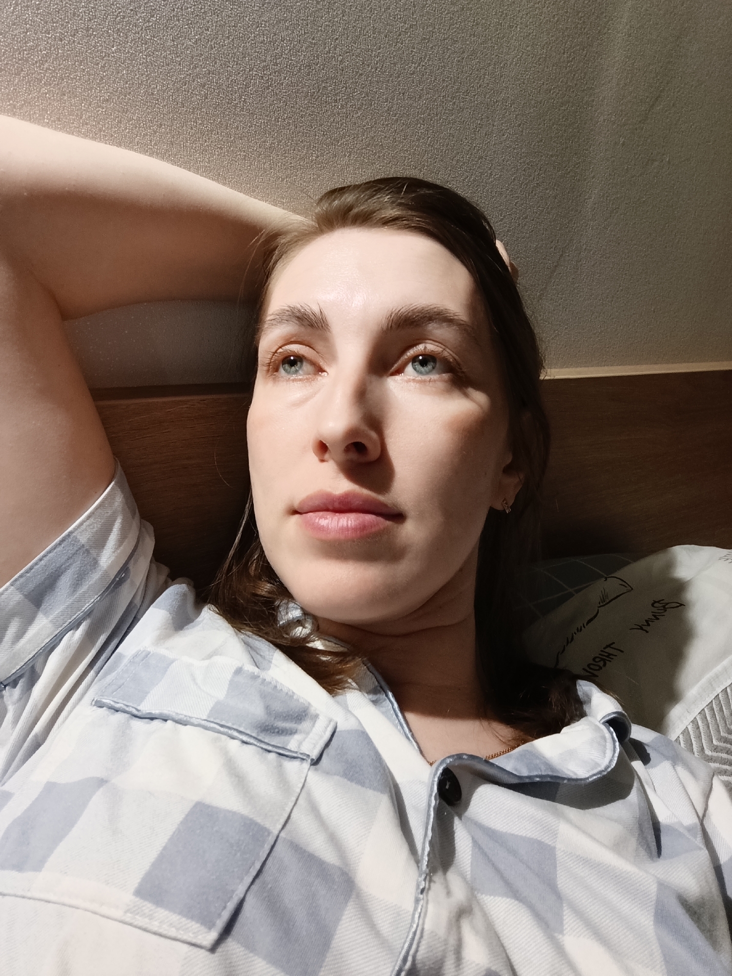 A fair-skinned woman lies in bed, wearing a blue and white plaid pajama top. Her arm is raised behind her head, and she is looking upwards and to the left with blue eyes. Her brown hair is partially pulled back. A pillow with writing in a language other than English is visible behind her head. The background includes a dark wooden headboard and a textured wall. The scene is likely indoors. There is no information available to confirm the location of the image is in Chaikovsky, Russia.