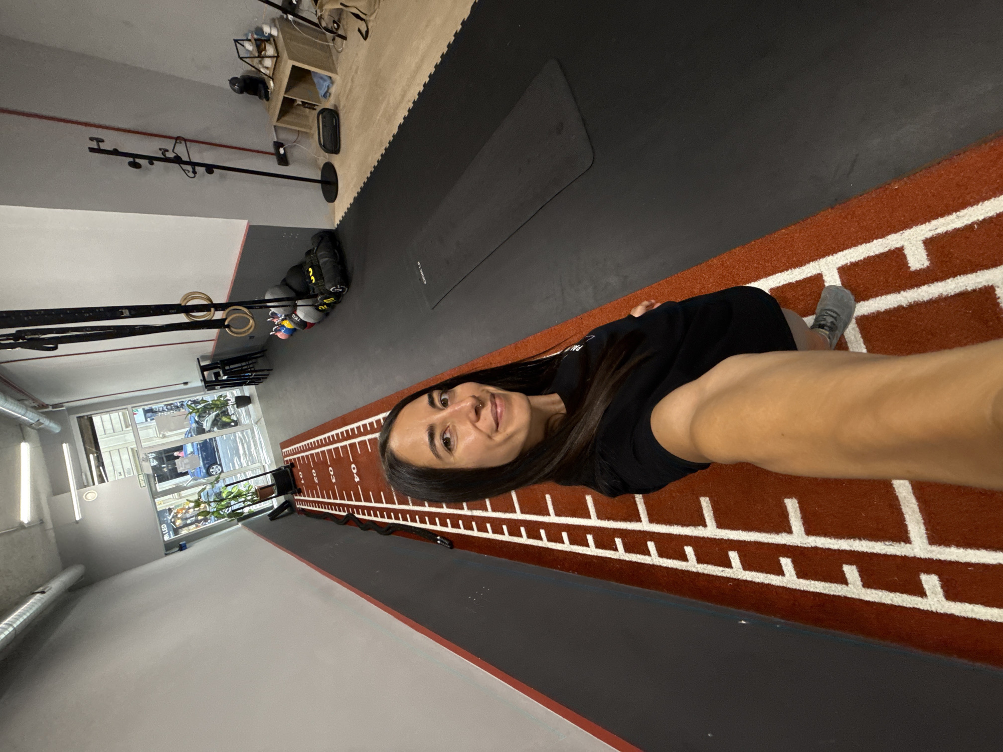 The image depicts a young woman in a gym, likely in Barcelona, Spain. She is taking a selfie, with her face positioned in the upper right quadrant of the frame. The woman has long, dark hair and is wearing a black top. Her left arm is extended, holding the camera, and her right arm is visible, resting on a textured, reddish-brown mat that appears to be part of a gym floor. This mat has white markings resembling a measuring tape or track, with numbers such as "0", "1", "2", "3", and "4" visible.
The setting is a gym with various equipment. In the background, to the left, there is a rack with gym rings and straps. Further back, there is a wooden shelf unit with items on it, and a black exercise mat is laid out on a dark floor. The gym appears to have large windows on one wall, offering a view of a street scene with buildings, plants, and vehicles, suggesting it's daytime. The overall lighting suggests an indoor environment, possibly with natural light coming from the windows. The woman's expression is neutral and directed towards the camera, indicating she is aware of being photographed. The presence of gym equipment and the mat with markings clearly identifies the activity as related to fitness or exercise.