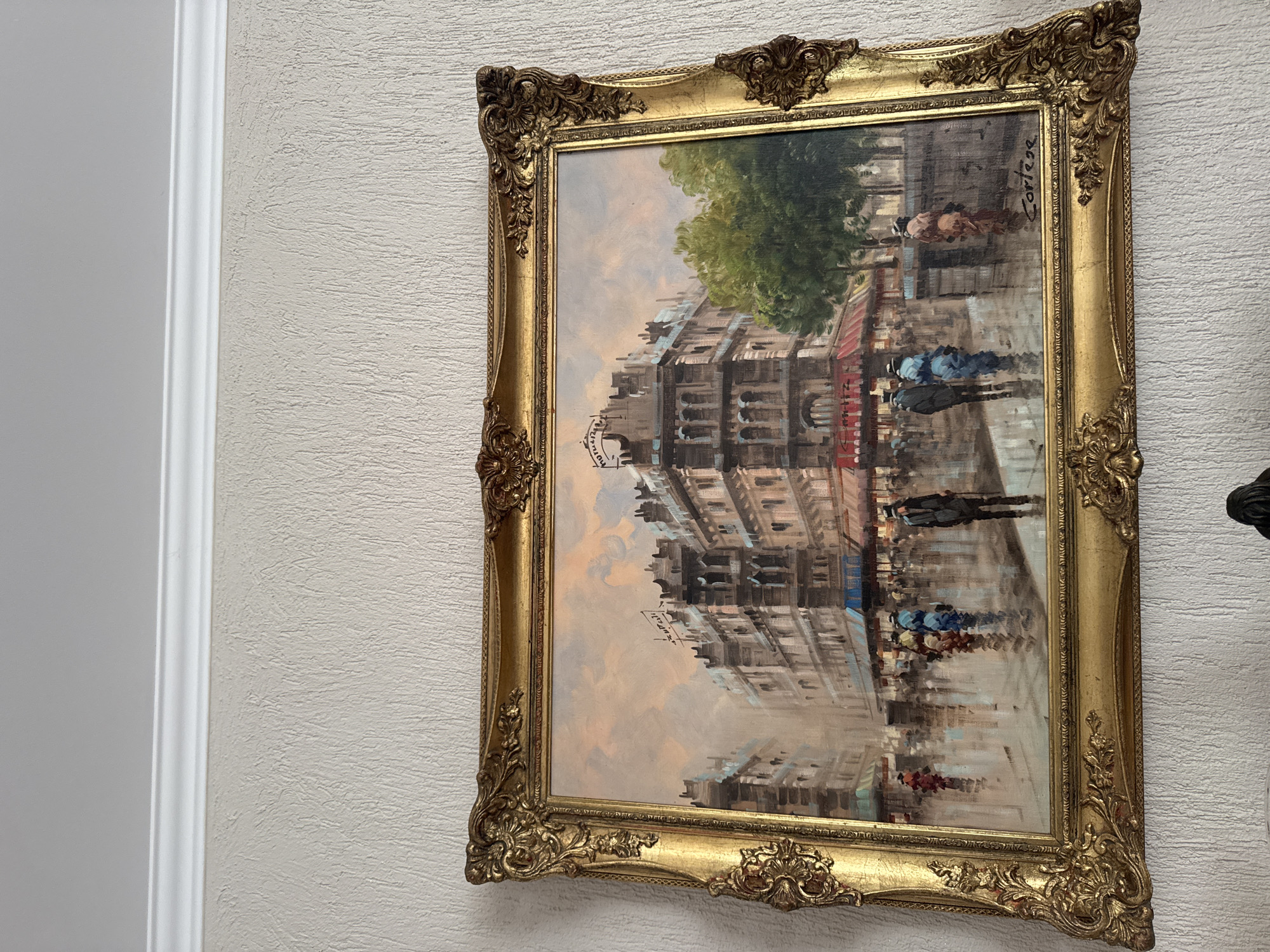 An oil painting of a Parisian street scene is displayed in an ornate gold frame hanging on a light-colored, textured wall next to a white wall panel. The painting depicts a street scene with buildings, a tree, and people with umbrellas, rendered in an impressionistic style with visible brushstrokes and reflections suggesting wet pavement. The painting is signed "Cortese" in the upper right corner. The scene suggests a rainy day in a European city.