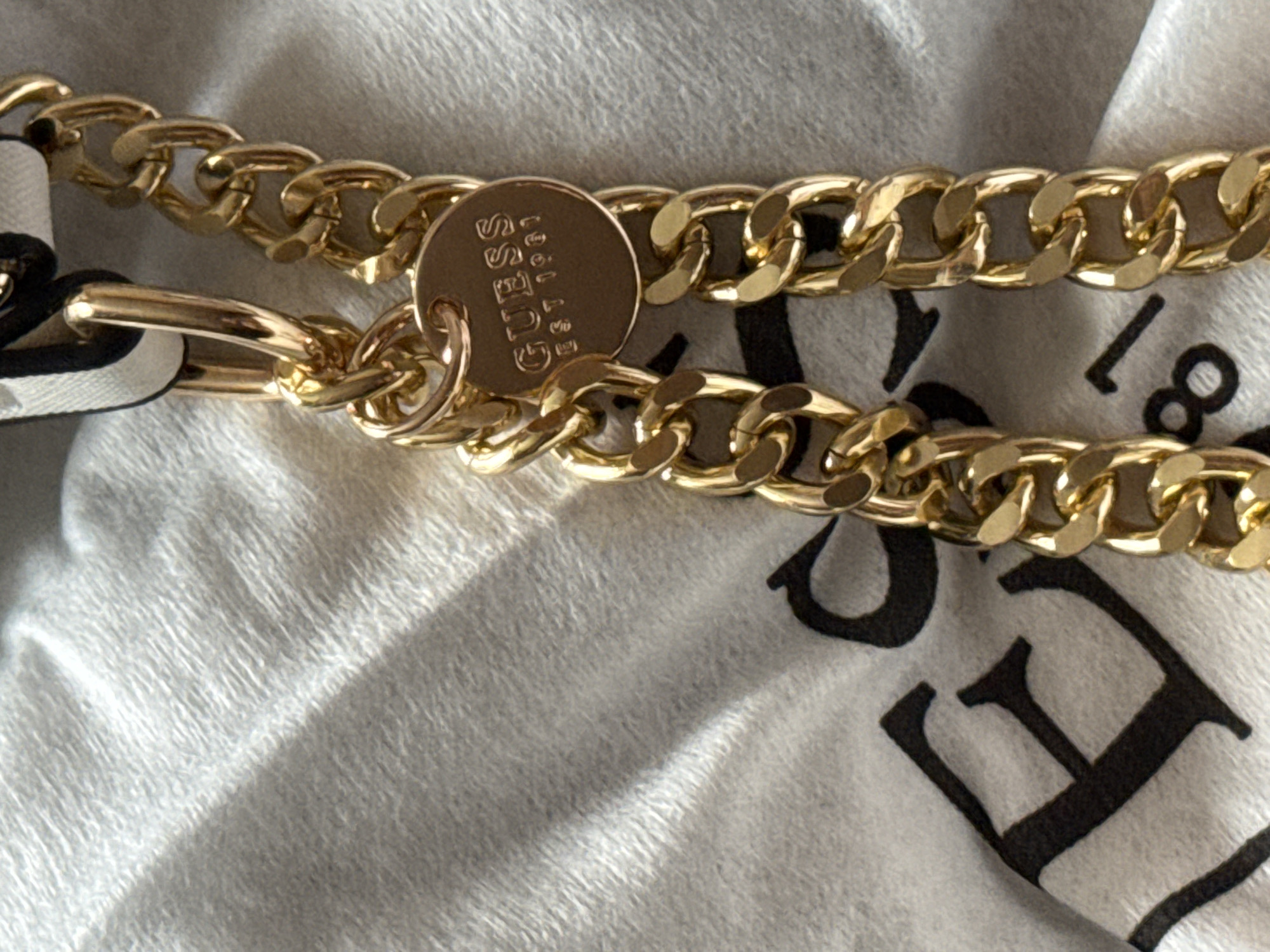 The media file shows a close-up of a gold-colored chain against a light-colored, textured fabric that appears to be a soft cloth or material. The chain is made of thick, interlocking links. Attached to the chain is a circular metal charm, also gold-colored, with embossed text. The text on the charm is partially visible and reads "GUESS" and "Est. 1981". Part of another chain is visible below the main one. The fabric has a subtle pattern or texture that creates shadows and highlights, suggesting a light source is present. In the background, on the fabric, there are large black letters that appear to be part of a logo or design, with some letters like "E" and "I" being partially discernible. No people, animals, or specific outdoor location cues are visible. The image focuses on the detail and texture of the jewelry and the fabric.