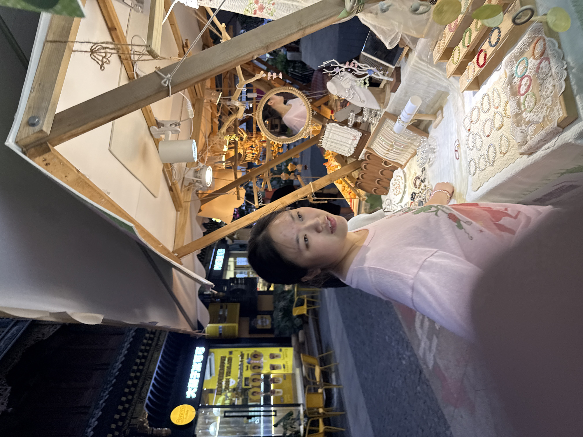 A young East Asian woman with dark hair pulled back is standing at a market stall in Yuzhong District, China. She is wearing a light pink t-shirt with a graphic design. The stall is wooden and displays various handcrafted jewelry, including necklaces, bracelets, and earrings, arranged on a white lace tablecloth and wooden stands. A round mirror with a decorative frame hangs on the stall, reflecting the woman's profile. In the background, a building with traditional Chinese architecture is visible, along with a brightly lit shop displaying drinks and seating areas. The street is paved, and the scene appears to be at dusk or night, with artificial lighting illuminating the stall and surrounding area.