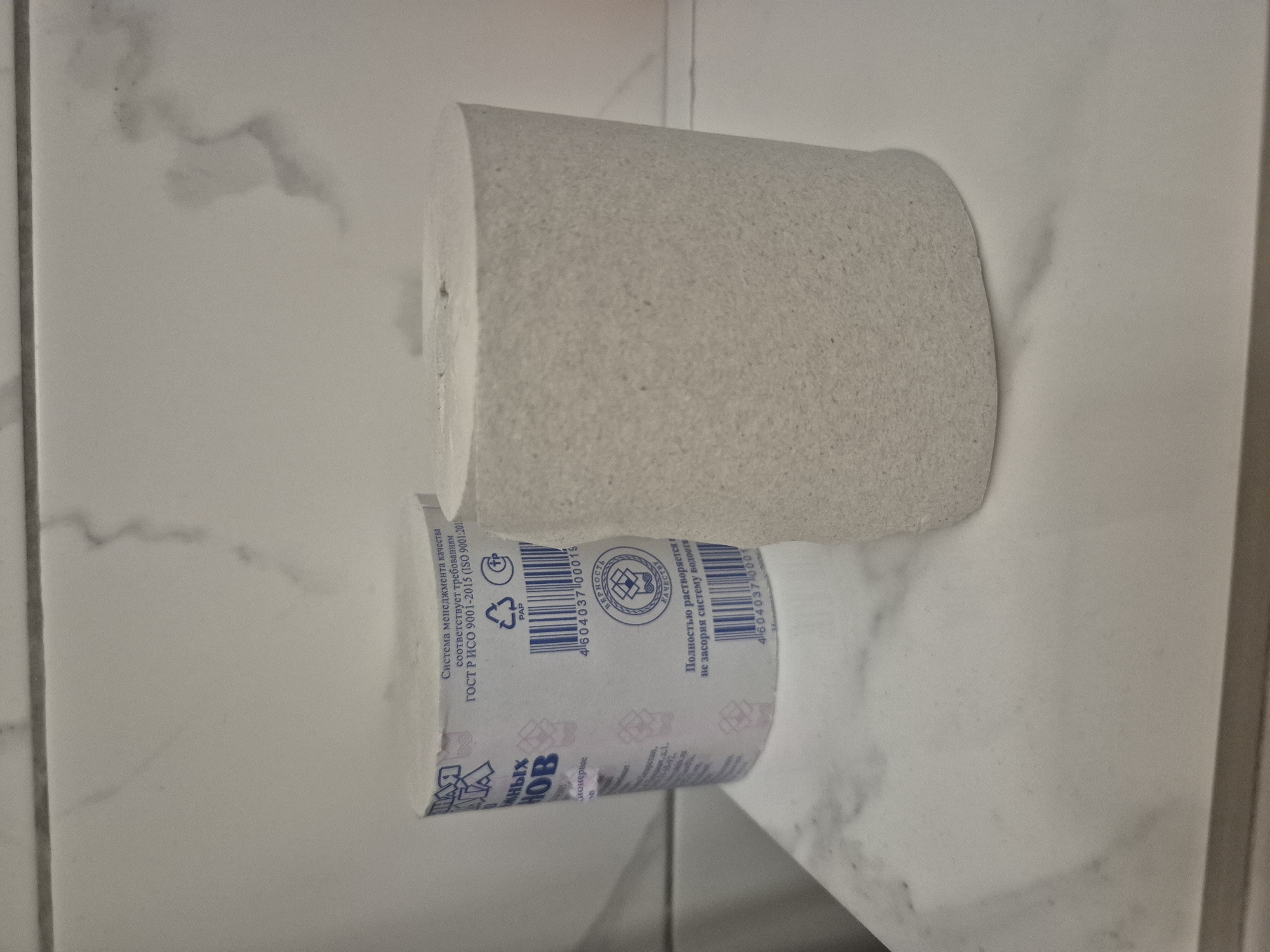 The image shows two rolls of toilet paper on a white marble surface. The roll on the left has a blue and white label with Russian text and a barcode. The roll on the right is plain and appears to be made of recycled paper. The background is a white tiled wall with subtle gray veining. There are no people or animals present in the image, and no discernible location in Astana, Kazakhstan can be identified.