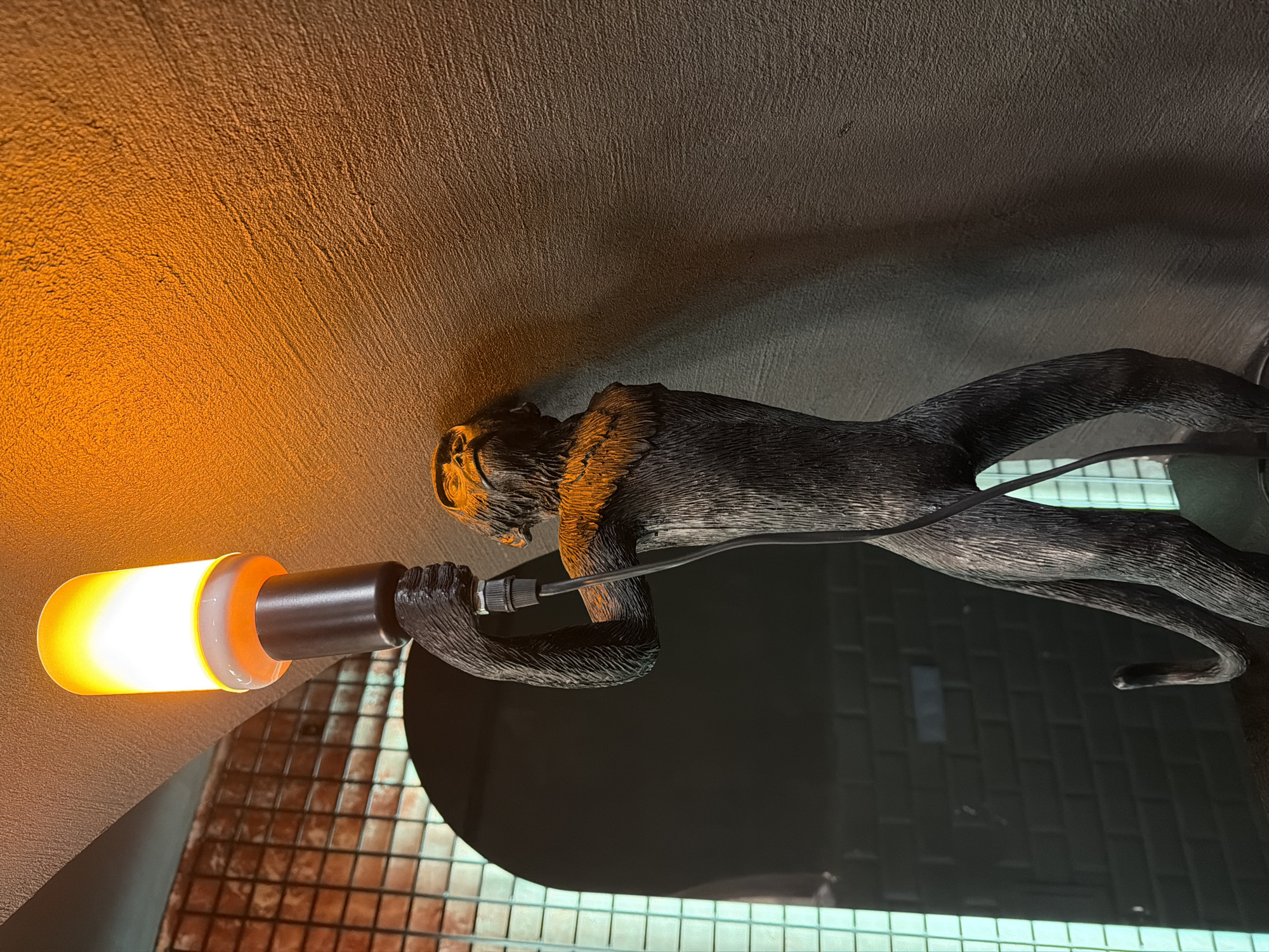 A sideways image depicts a decorative light fixture featuring a monkey statuette holding a lightbulb. The monkey is dark brown or black, with textural details suggesting it's made of wood or a similar material. It lies on a ceiling or wall, clutching a cylindrical lightbulb with a bright yellow glow at one end. The lightbulb is white and translucent near its base. A black cord extends from the back of the monkey’s head, suggesting it's wired to the lightbulb. The background consists of a textured surface, possibly a painted ceiling or wall, with an orange tint from the light. In the lower portion of the image, a tiled wall is visible. The overall impression is that of a quirky, artistic light fixture installed in a room. There is no information available from the image to identify the location in Izhevsk, Russia, or to identify any people, actions, or interactions.