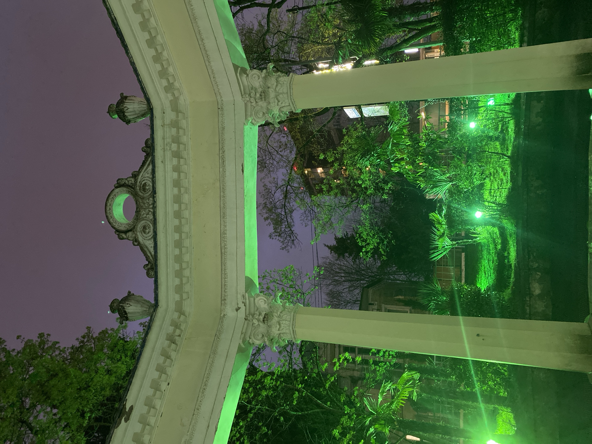 The image shows an architectural structure in Sochi, Russia. The structure is made of columns and an arch, likely located in a park or garden. The architecture is ornate, with decorative carvings. The scene is lit with green lights, which illuminate the trees and foliage. The sky is dusky, suggesting it is evening or night.