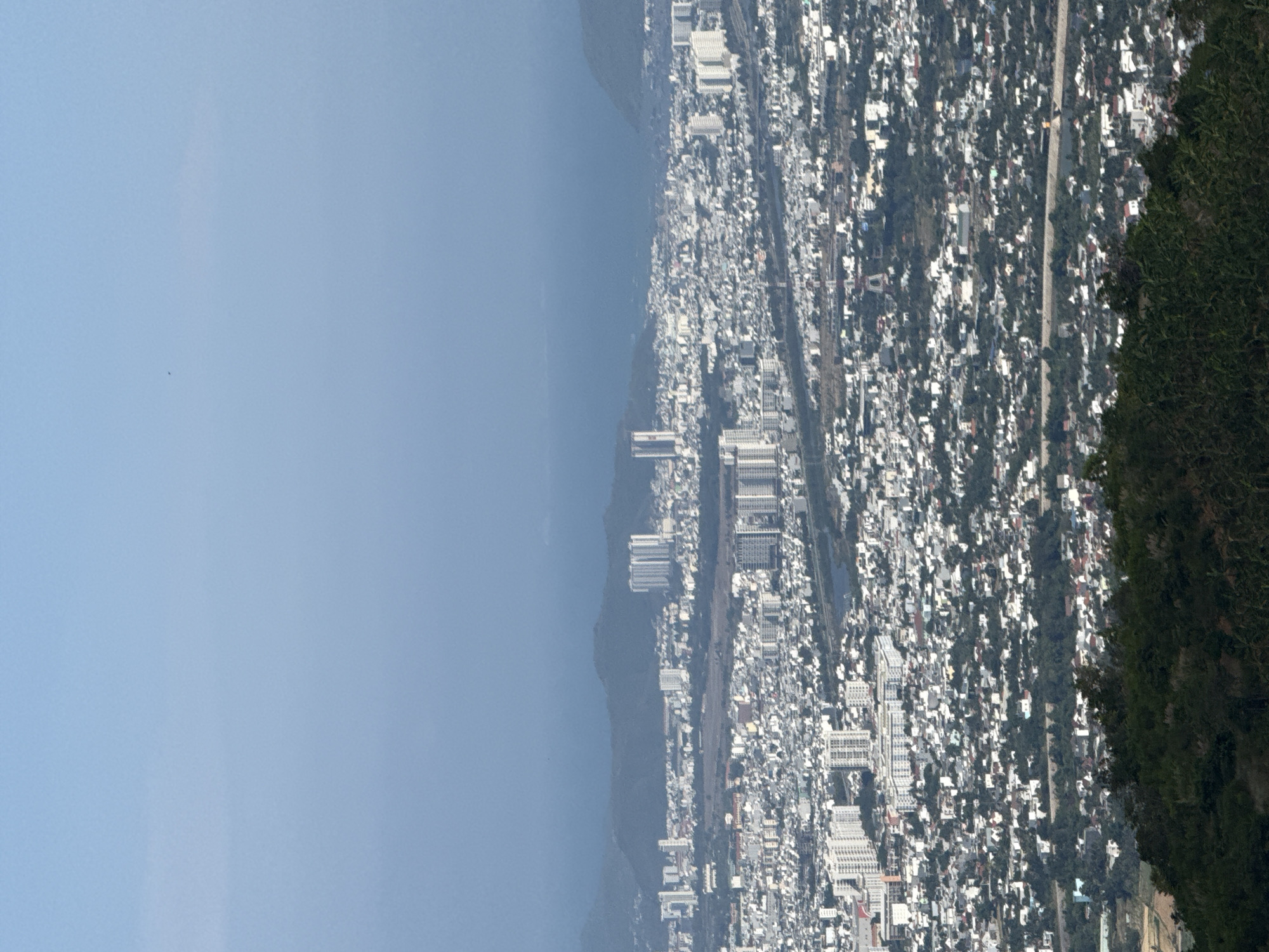 This image presents an elevated panoramic view of a coastal city, identified as Tay Nha Trang Ward, Vietnam. The scene is dominated by an expansive, clear blue sky occupying the upper left portion. Below the sky lies a vast, calm expanse of blue sea, stretching horizontally across the mid-ground.
Beyond the immediate coastline, a long, low mountain range appears slightly hazy in the distance, separating the sea from the urban area in some sections. The main subject is a densely populated city, sprawling across the landscape to the right of the sea and distant mountains. This urban area features a mix of numerous white and light-colored buildings, ranging from low-rise structures to several multi-story high-rise buildings scattered throughout. Roads and infrastructure are visible winding through the city.
On the far right of the frame, a steep, dark green, forested hill or mountain dominates the foreground, partially obscuring the city below and providing a natural boundary.
The time of day appears to be mid-day or early afternoon, characterized by bright, even sunlight and a clear sky, indicating good weather conditions. No people or specific activities are discernible, as it is a wide landscape shot. There is no visible text in the image.