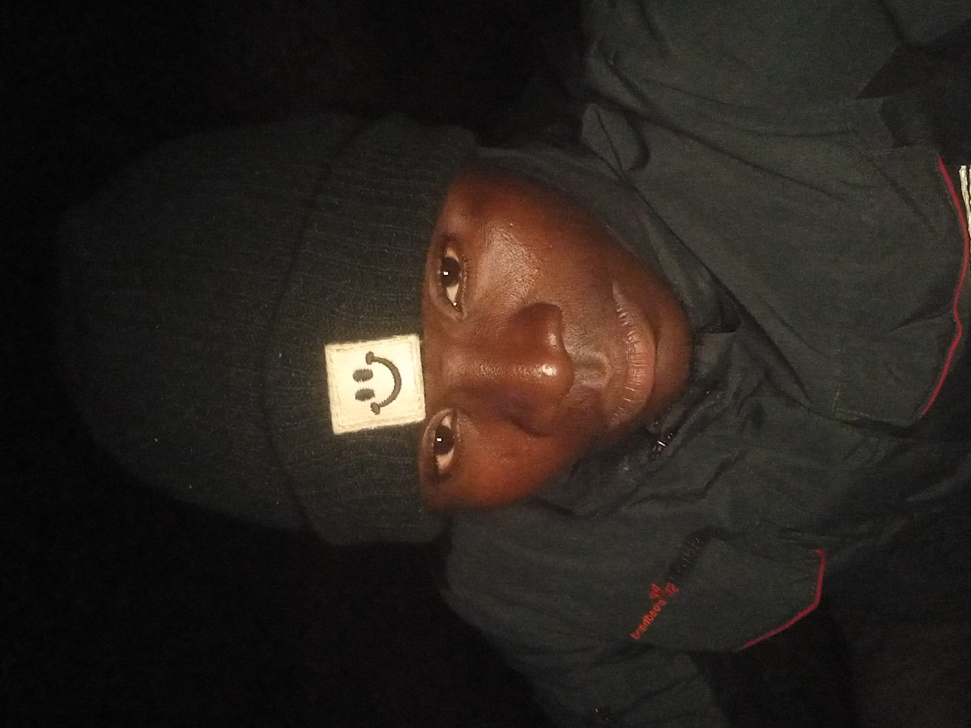 A person is wearing a black beanie with a yellow smiley face patch and a black jacket. The person is looking at the camera. The image appears to be a selfie taken at night. The location in Kwang, Nigeria is not evident from the image.