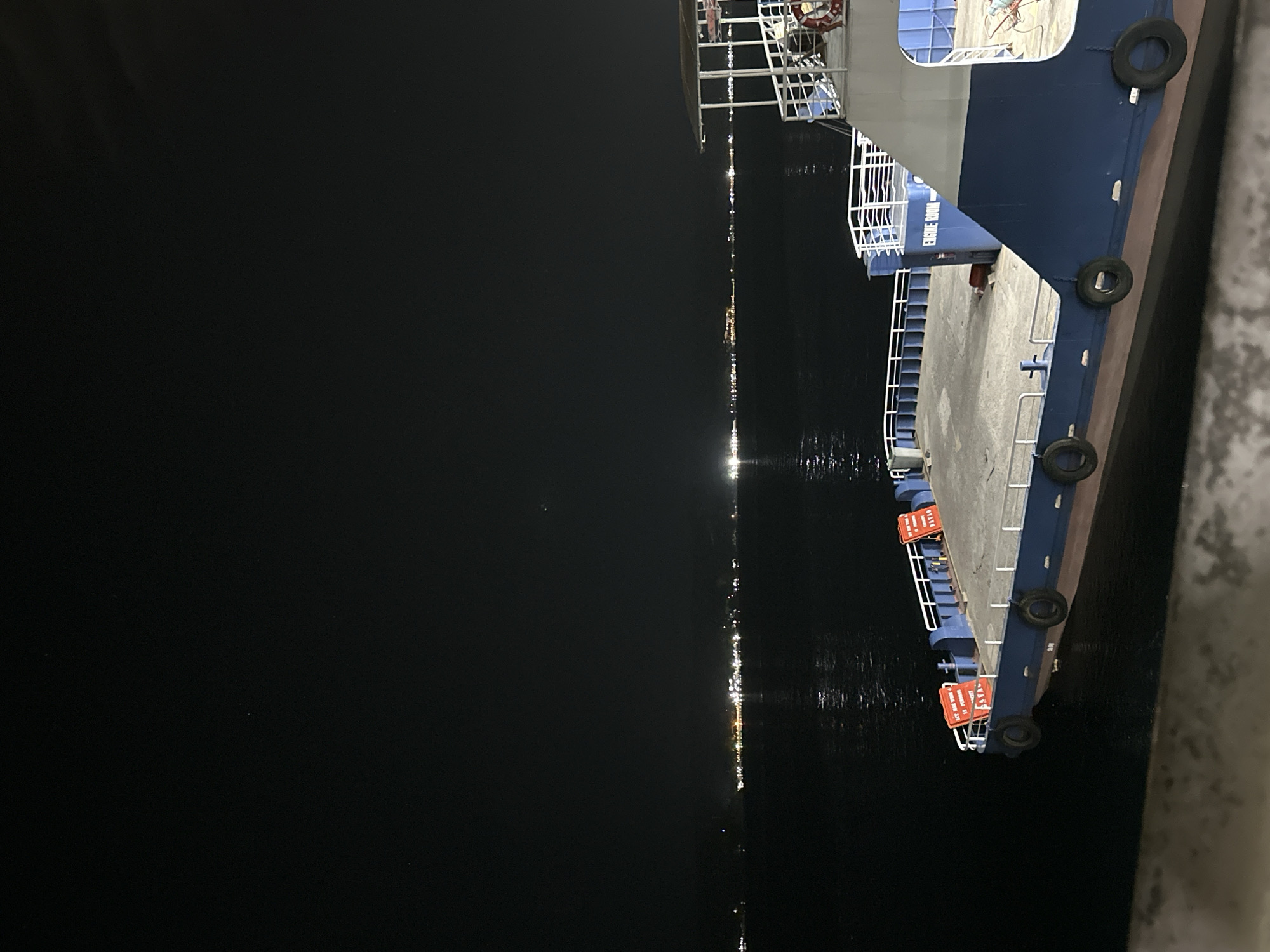 This nighttime image depicts a large blue and white vessel moored at a pier, likely in Davao City, Philippines. The boat occupies the right side of the frame, showcasing its sturdy blue hull, white railings, and several black rubber fenders hanging along its side, indicating it is securely docked.
On the boat's structure, part of a white cabin or superstructure is visible, with text that appears to read "ENGINE ROOM". Along the boat's railing, several rectangular orange signs are attached. One sign clearly shows the text "NO SMOKING", while another, partially obscured, appears to have "ICE BOX" and possibly "PATIO" or similar words.
To the left of the boat, a vast, dark body of water stretches into the distance. The water's surface is calm, reflecting a long, shimmering line of bright lights from a distant shore or other vessels, creating a striking contrast against the darkness. The sky is completely black, indicating a clear night with no visible stars or moon. The scene is quiet and still, emphasizing the artificial lights against the natural darkness of the night and water. No people are visible, and the scene suggests a quiet moment at a busy port.
