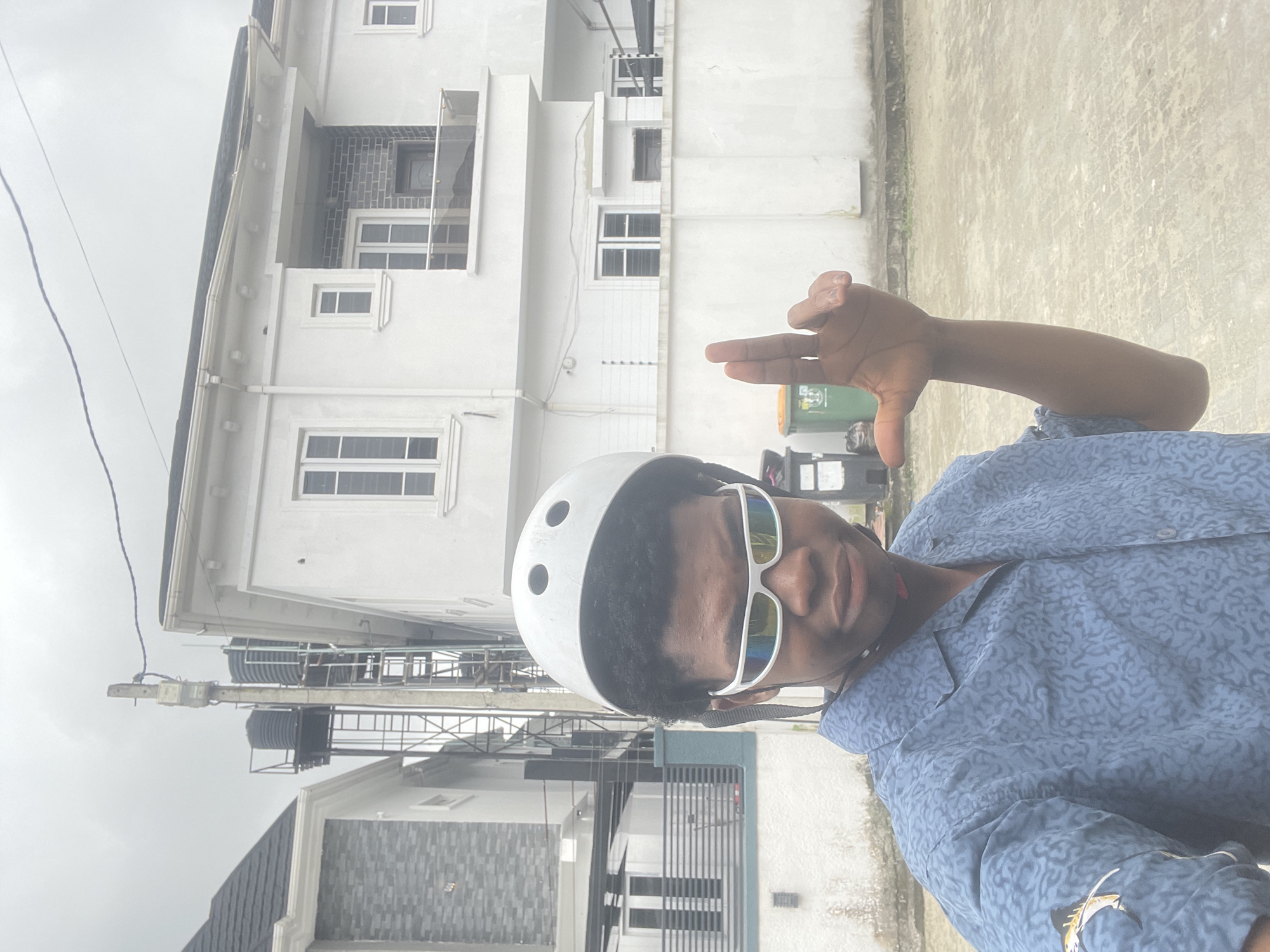 In Ado, Nigeria, under an overcast sky, a young Black male stands on a paved surface. He wears a white helmet and reflective white sunglasses. His blue patterned shirt features a light-colored fish graphic on the left sleeve. His right hand is raised, with index and middle fingers extended in a 'V' shape. Behind him, modern white houses with dark-trimmed windows and balconies are visible. A concrete pole supporting water tanks and electrical cables stands between the houses. Green and black trash bins are situated near a white concrete wall.