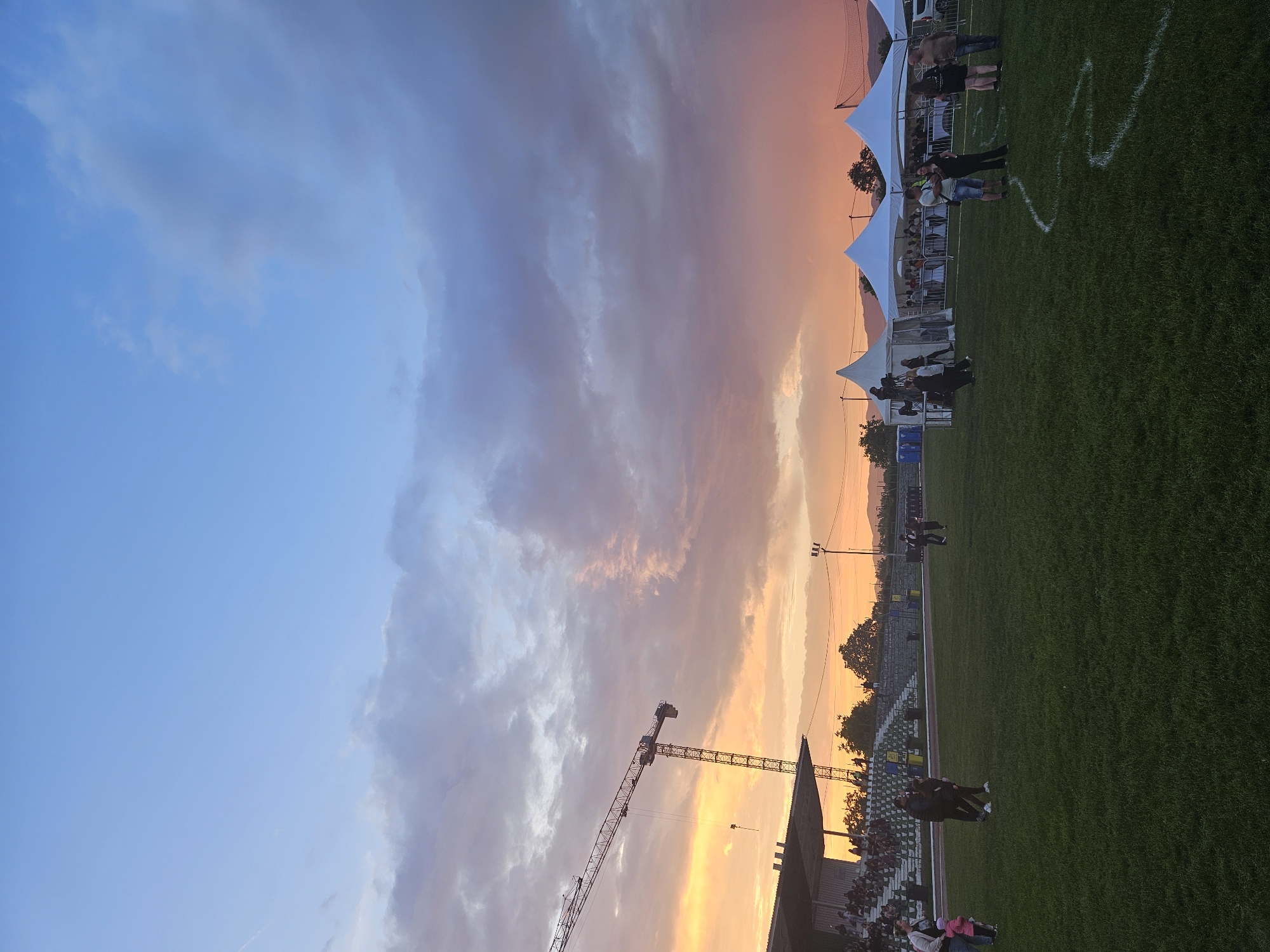The image captures an outdoor event on a grassy field in Elin Pelin, Bulgaria, during a sunset. The sky displays a gradient of colors, from light blue at the top fading into orange, pink, and purple hues closer to the horizon, with various cloud formations. A tall construction crane is prominently silhouetted against the vibrant sky in the upper left-center. Below the crane and to the left, a stadium grandstand, featuring multiple rows of seating and a roof structure, is partially filled with spectators.
On the right side of the grassy field, a series of white, peaked event tents are erected, with numerous individuals gathered around and inside them. Two blue portable toilets are visible behind one of the tents on the left side of the tent cluster. A tall light pole and overhead wires are present in the mid-right background, and a section of a volleyball net is visible in the far right distance. Distant mountains or hills form a dark silhouette against the horizon.
In the foreground and mid-ground, multiple people are present on the grass. On the left, an adult is standing with a small child near the grandstand. On the right, groups of adults are conversing and moving around the event tents. A white, wavy line is painted on the grass in the lower right portion of the field. The overall scene indicates a public gathering or festival occurring at a sports complex during the late evening.