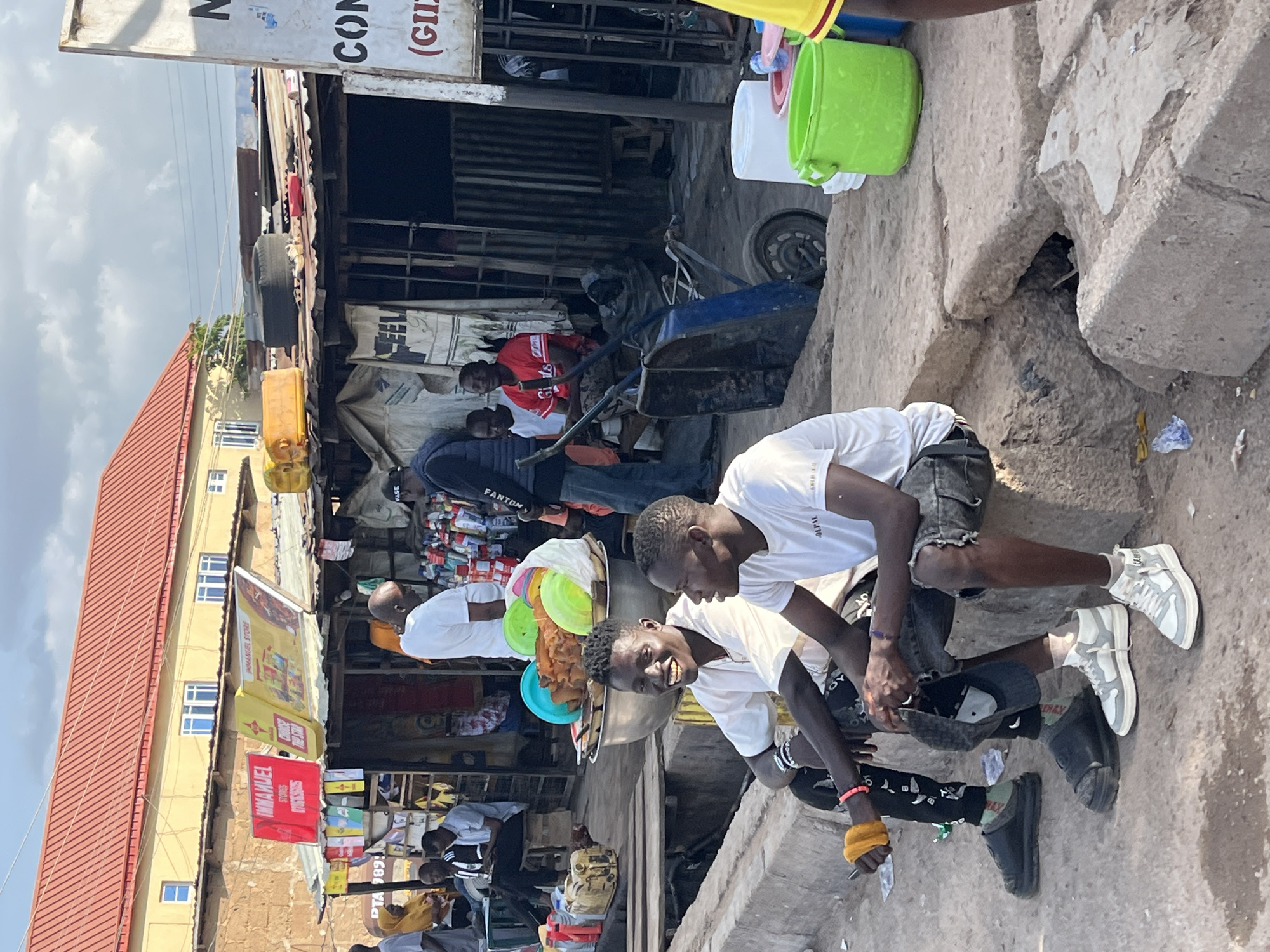 This image depicts a street scene in Gura Top, Nigeria, with an emphasis on a marketplace or roadside stalls. Two young men are prominently seated on a concrete curb in the foreground, facing towards the right of the frame. The man on the left is wearing a white t-shirt, black pants with white patterns, and black slides. He is holding a yellow object in his left hand. The man on the right is wearing a white t-shirt and dark denim shorts, with white sneakers. Both men are looking to their right and appear to be engaged in conversation or observing something outside the frame.
Behind them, a bustling market environment unfolds. Several stalls with corrugated metal roofs are visible, displaying various goods. One stall has large signs with "IMMANUEL STORES" and "CHOOSE THE STAR" printed on them. Other stalls display an assortment of items, including what appears to be packaged goods, plastic containers, and perhaps food items. People are seen moving around and interacting within the market area. A cart with a blue frame is parked near one of the stalls.
In the background, a yellow building with a red roof stands, suggesting a more permanent structure adjacent to the market. The sky is overcast with clouds, indicating a daytime scene. The overall impression is one of a lively and active local commerce area.