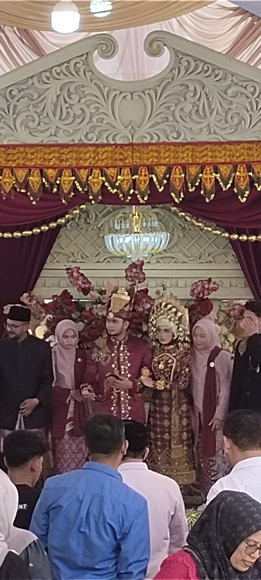 Captured in an unidentified location in the city of Unknown, Unknown, the photograph shows a wedding scene featuring the bride and groom standing under an ornate architectural structure adorned with burgundy drapes, floral arrangements, and a chandelier. The groom is dressed in a maroon suit with intricate gold detailing, while the bride wears a traditional dress with a matching headdress. Flanking them are several individuals in formal attire, and in the foreground, there is a crowd of guests.