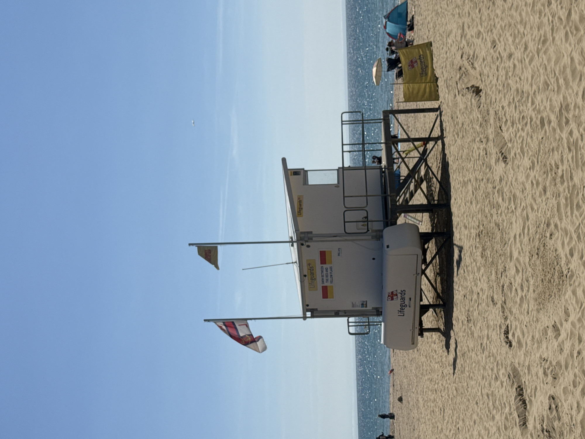This is a photograph of a lifeguard station on Bournemouth Beach in Bournemouth, United Kingdom. The station is a white, elevated structure with a roof and an enclosed cabin. There are two flags flying from poles next to the station: one is a red and white ensign, and the other is a yellow flag with a red cross. Text on the side of the station reads "Lifeguards" and "SWIM BETWEEN THE RED AND YELLOW FLAGS." A yellow windbreak with "Lifeguards" printed on it is positioned in front of the station. The beach is made of sand, and the sea is visible in the background. Several people are present on the beach, some sitting under umbrellas and others walking. There are also a couple of beach tents visible. The sky is clear and blue.