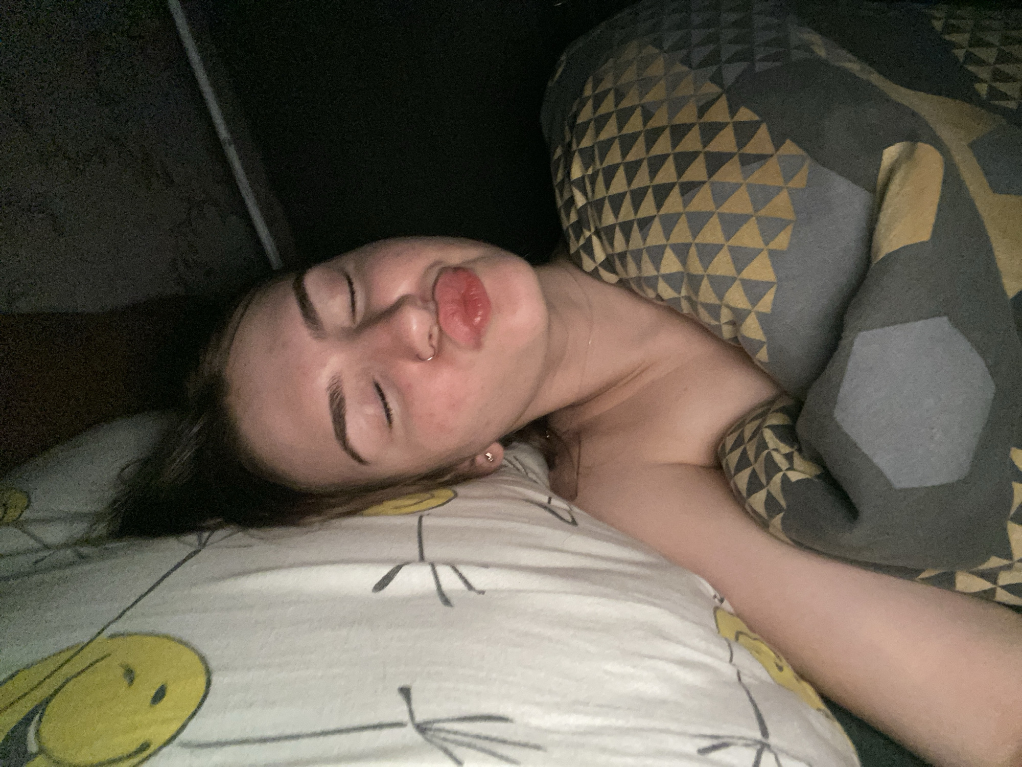 A young woman with fair skin and dark eyebrows is lying down, facing upwards, with her eyes closed and her lips pursed. She is wearing a nose ring. She is covered by a grey and yellow patterned blanket and rests her head on a white pillow with a yellow smiley face pattern. The environment appears to be a bedroom, with dark, out-of-focus surroundings. There is no discernible evidence of the city of Dyakovo, Russia, in the image.