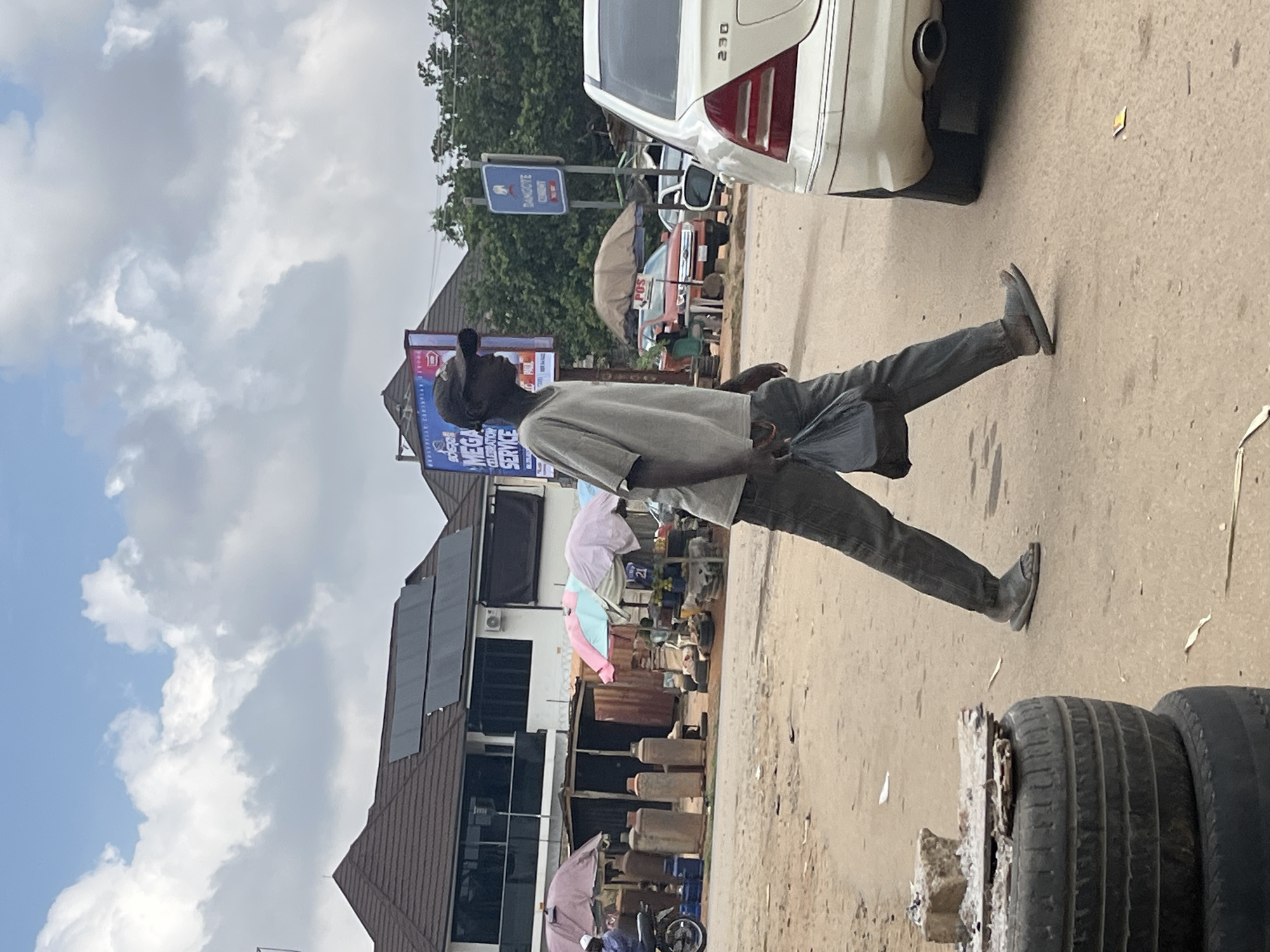 A young man walks across a dusty street in Abuja, Nigeria, carrying a black plastic bag. He is wearing a light gray t-shirt, dark pants, and sandals. In the background, there is a modern building with solar panels on the roof, a market stall with umbrellas and propane tanks, and a white Mercedes-Benz sedan. A blue sign for "Damcote Cement" is also visible. The sky is partly cloudy. The overall scene depicts a street view in a commercial area.