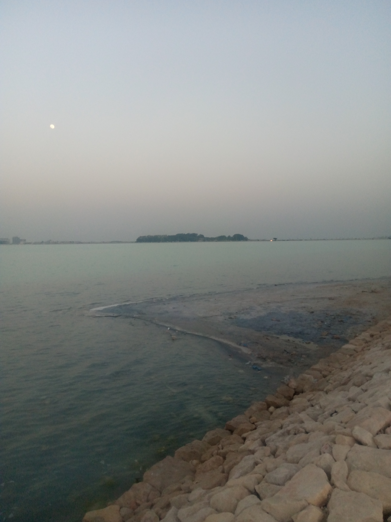 The image depicts a coastal scene at dusk in Al Khobar, Saudi Arabia. A body of water, possibly the Arabian Gulf, stretches towards the horizon under a pale, overcast sky with a faint moon visible. A small, vegetated island is visible in the distance. The shoreline is reinforced with large, light-colored rocks. The water's edge meets a sandy or muddy transition zone. There are no people visible in the scene.