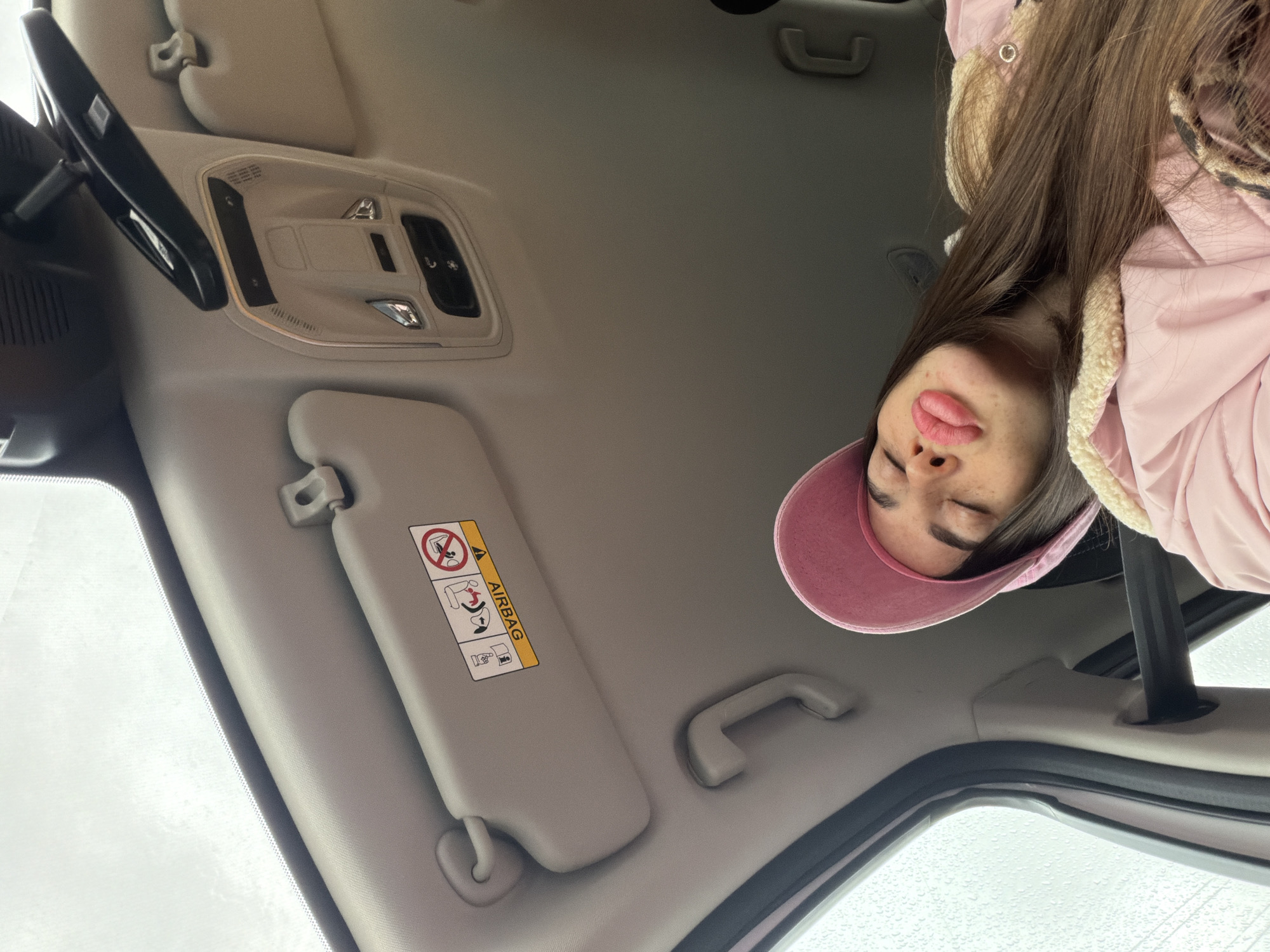 The image is taken from inside a car, looking upwards towards the ceiling and partially showing the person sitting in the car. The car's interior features a gray ceiling with a sun visor on the left, displaying an airbag warning label with pictograms. Above the sun visor, there's a ceiling light fixture. A grab handle is visible near the right edge of the frame, closer to the person. The person is wearing a pink jacket with a beige fur lining and a pink baseball cap. The person's face is partially visible, showing closed eyes and pursed lips. Through the sunroof, a view of a rainy environment is visible, suggesting it might be a car ride in inclement weather. The location is not discernible from the image.