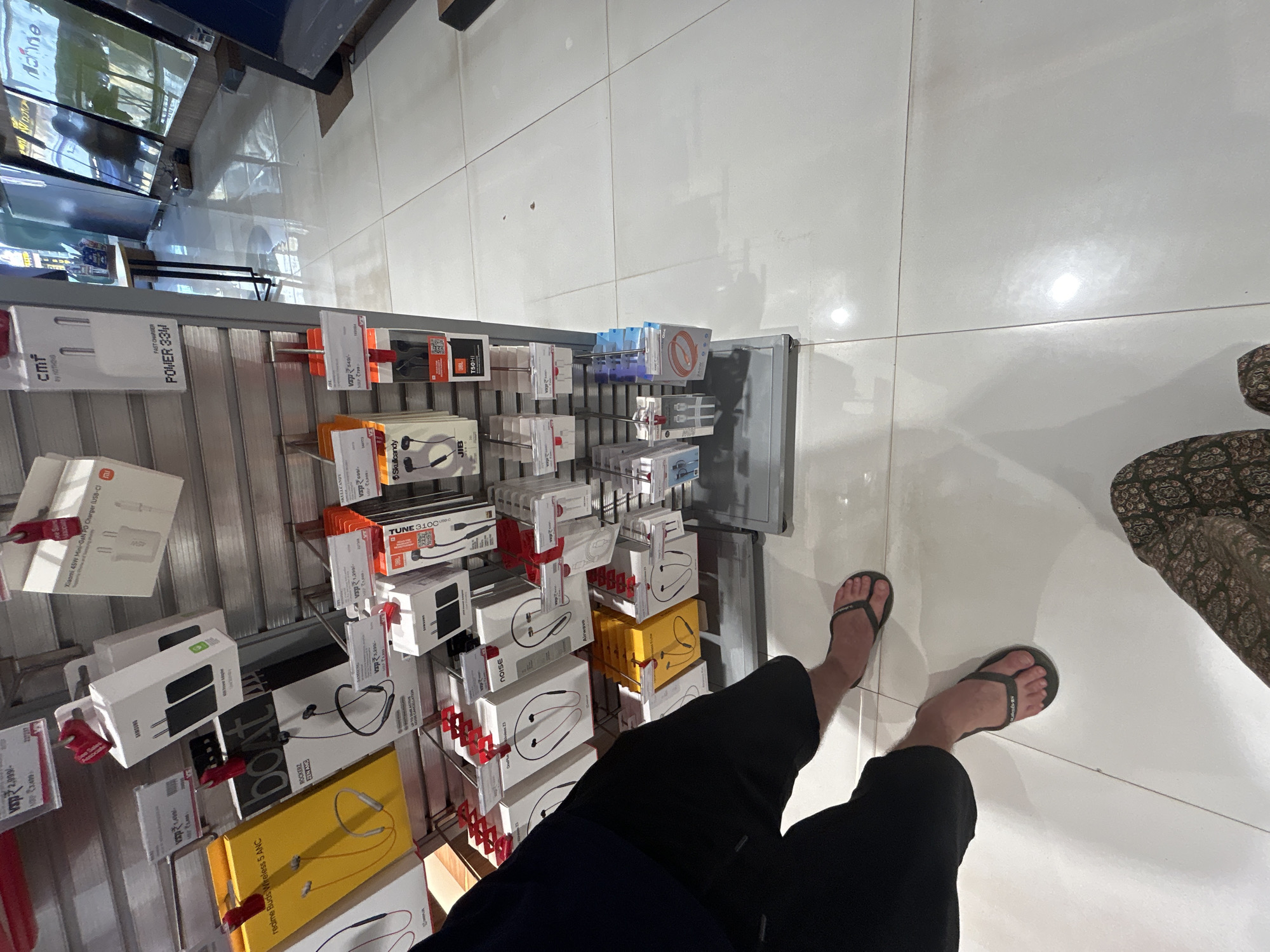 An eye-level shot shows a retail store display of electronic accessories. In the foreground, a person's legs wearing black shorts and black flip-flops are visible, standing on a white tiled floor. Another person's legs wearing patterned pants are also visible. The display racks are filled with various items such as earphones, charging adapters, and cables from brands like Samsung, Realme, Boat, JBL, and Noise. The store environment appears to be well-lit, with reflections visible on the floor. The overall impression is of a commercial setting, likely an electronics store in Mumbai, India, where customers are browsing products.