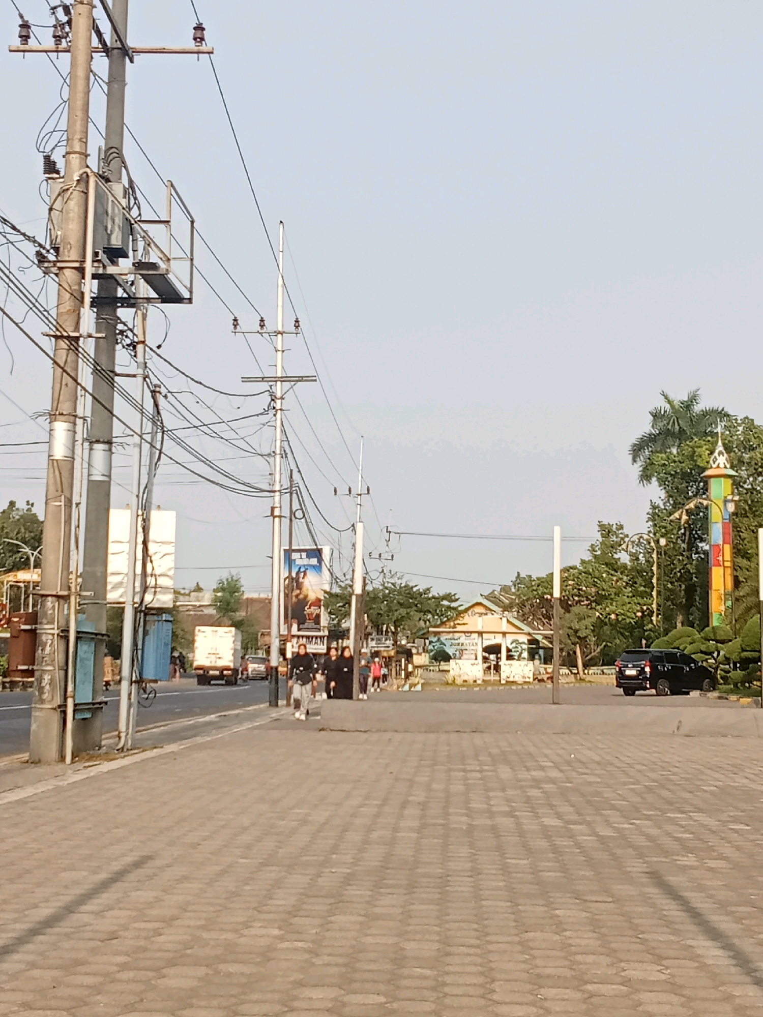 A sunlit, ground-level exterior shot captures a street scene in Kaliwungu, Indonesia, with a focus on a paved sidewalk in the foreground. A light pole with numerous wires and a transformer stands on the left. Across the street, a white truck and other vehicles are visible, along with pedestrians walking on the sidewalk. Further in the background, a building with a distinct roofline and a colorful, tower-like structure are present, surrounded by trees. A black SUV is parked on the right side of the road. The sky is clear and pale blue.